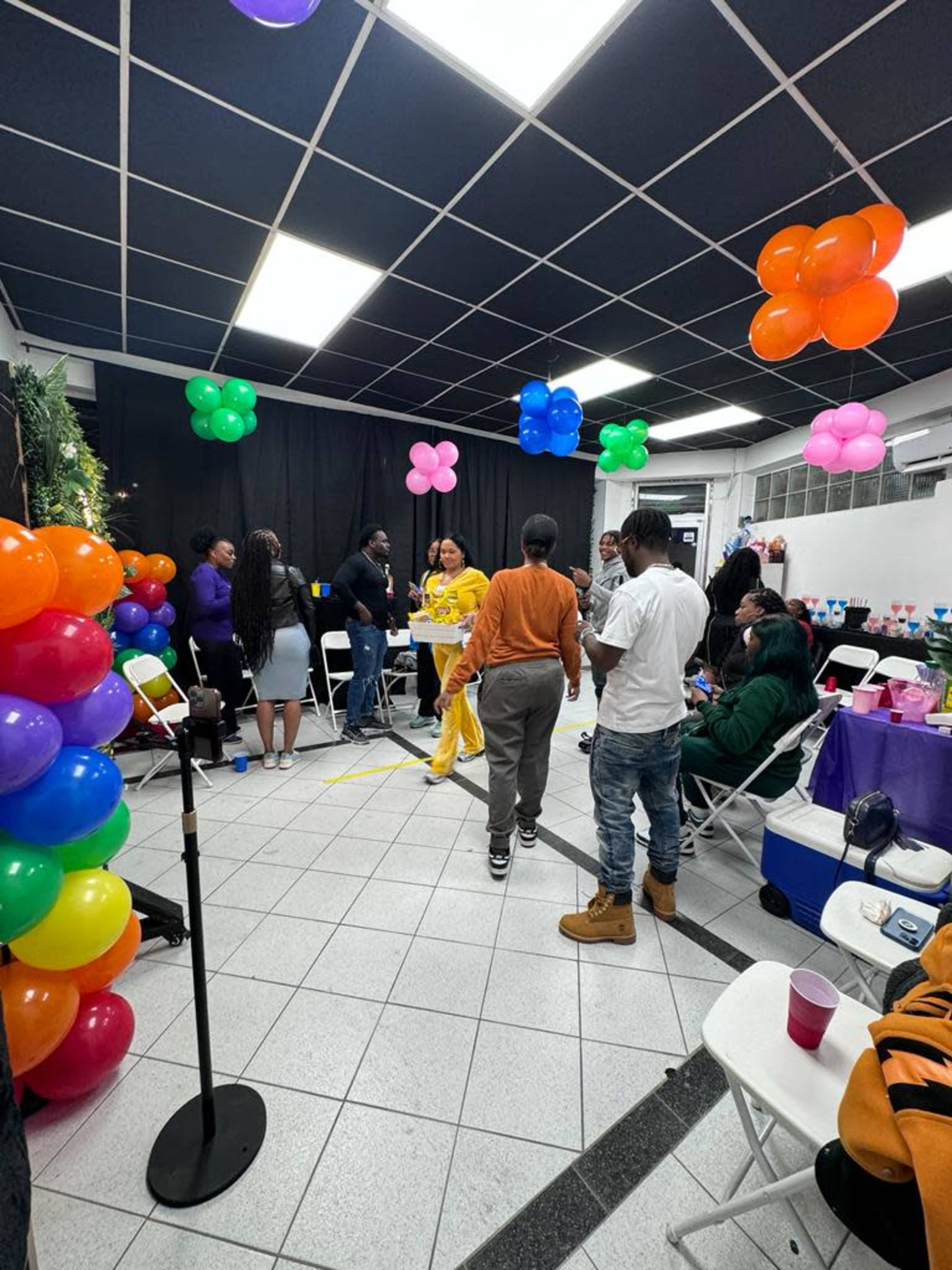 A group of people gathers in a decorated indoor space with colorful balloons and tables set up for an event.