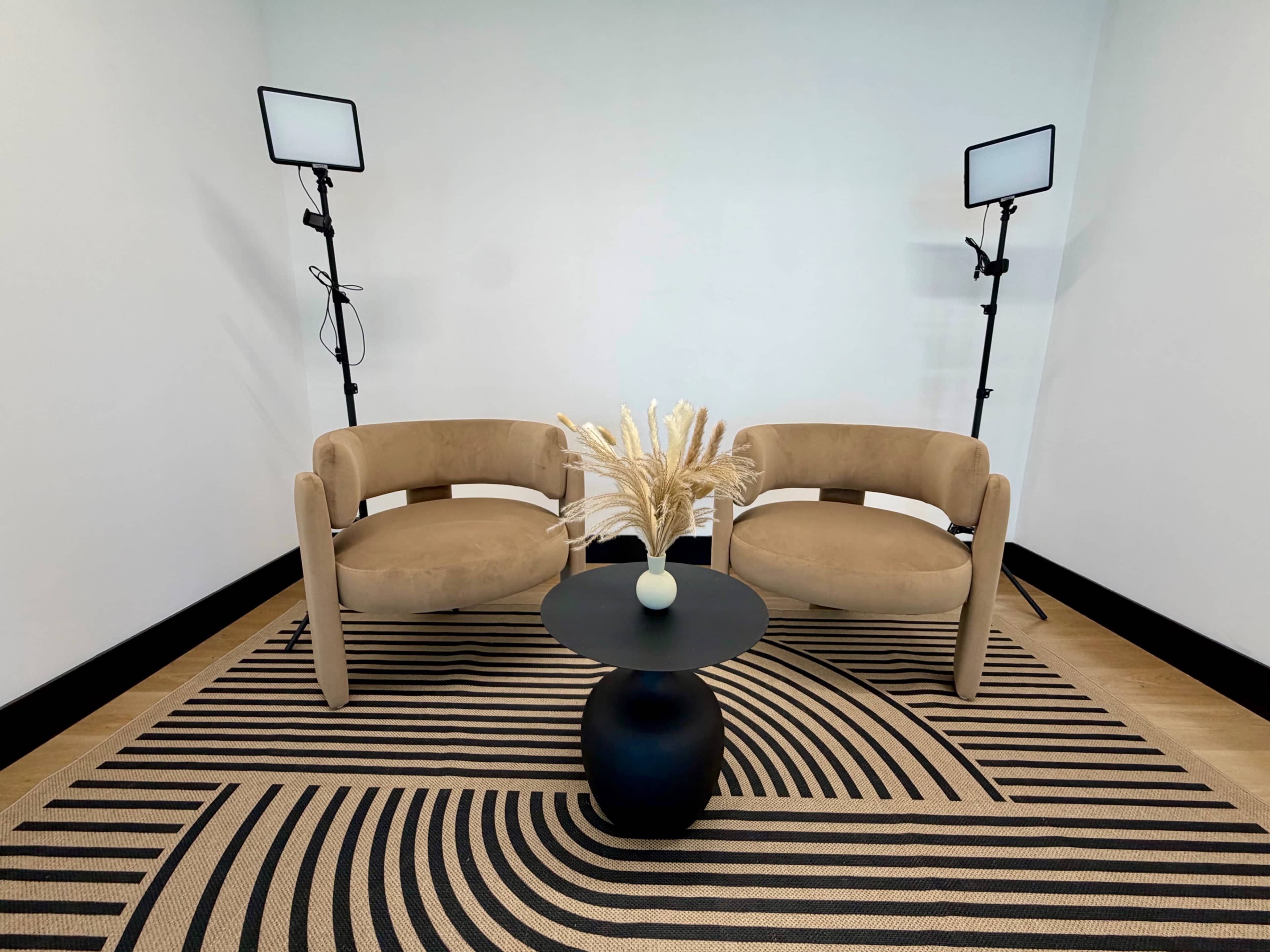 The image shows a minimalistic seating area with two beige chairs arranged around a black circular table, featuring a decorative vase and dried plants, set against a neutral wall and illuminated by two standing lights.