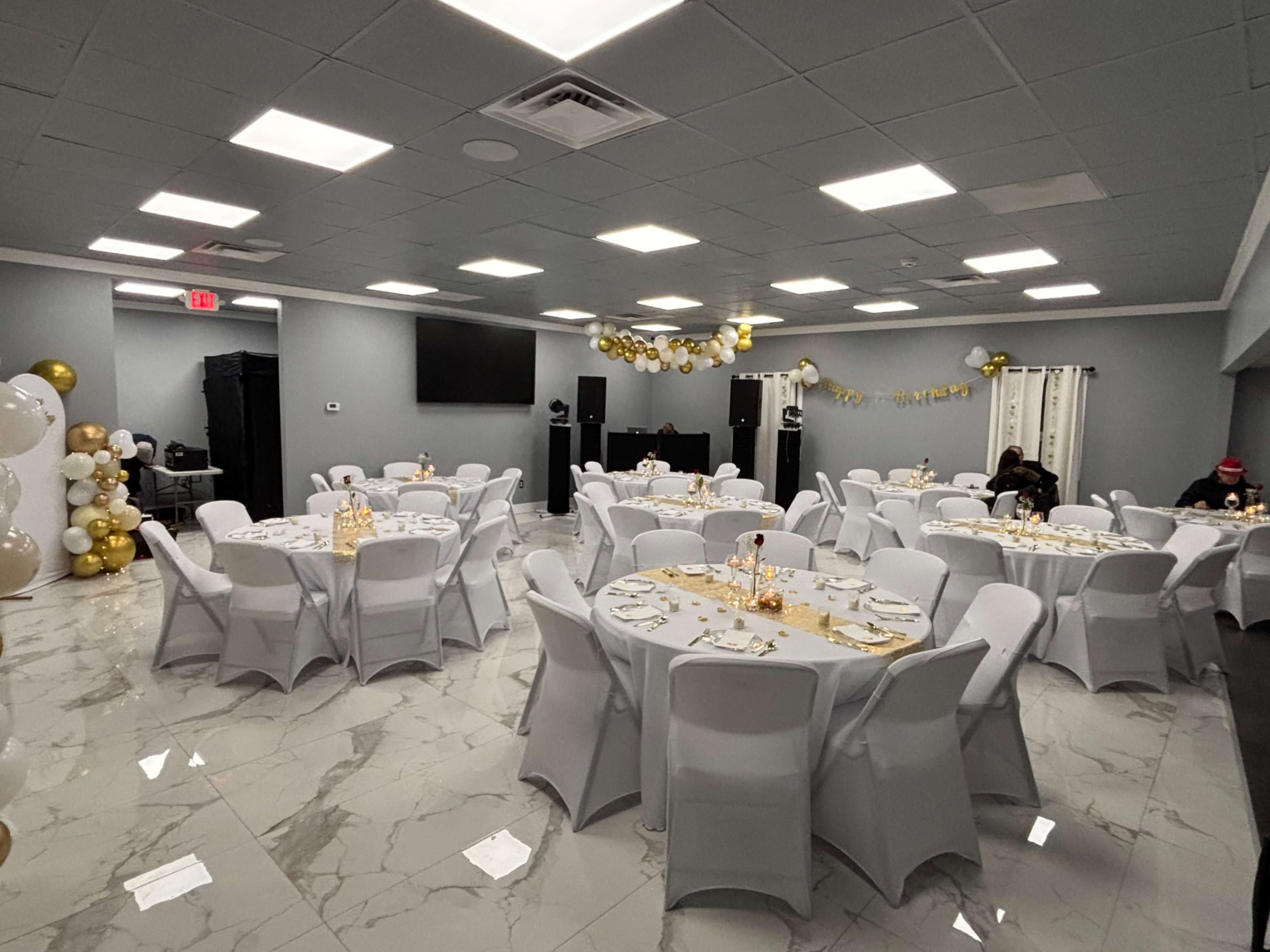 The image shows a banquet hall set up for an event, featuring round tables with white tablecloths, decorated with gold accents, and balloons arranged on the walls.
