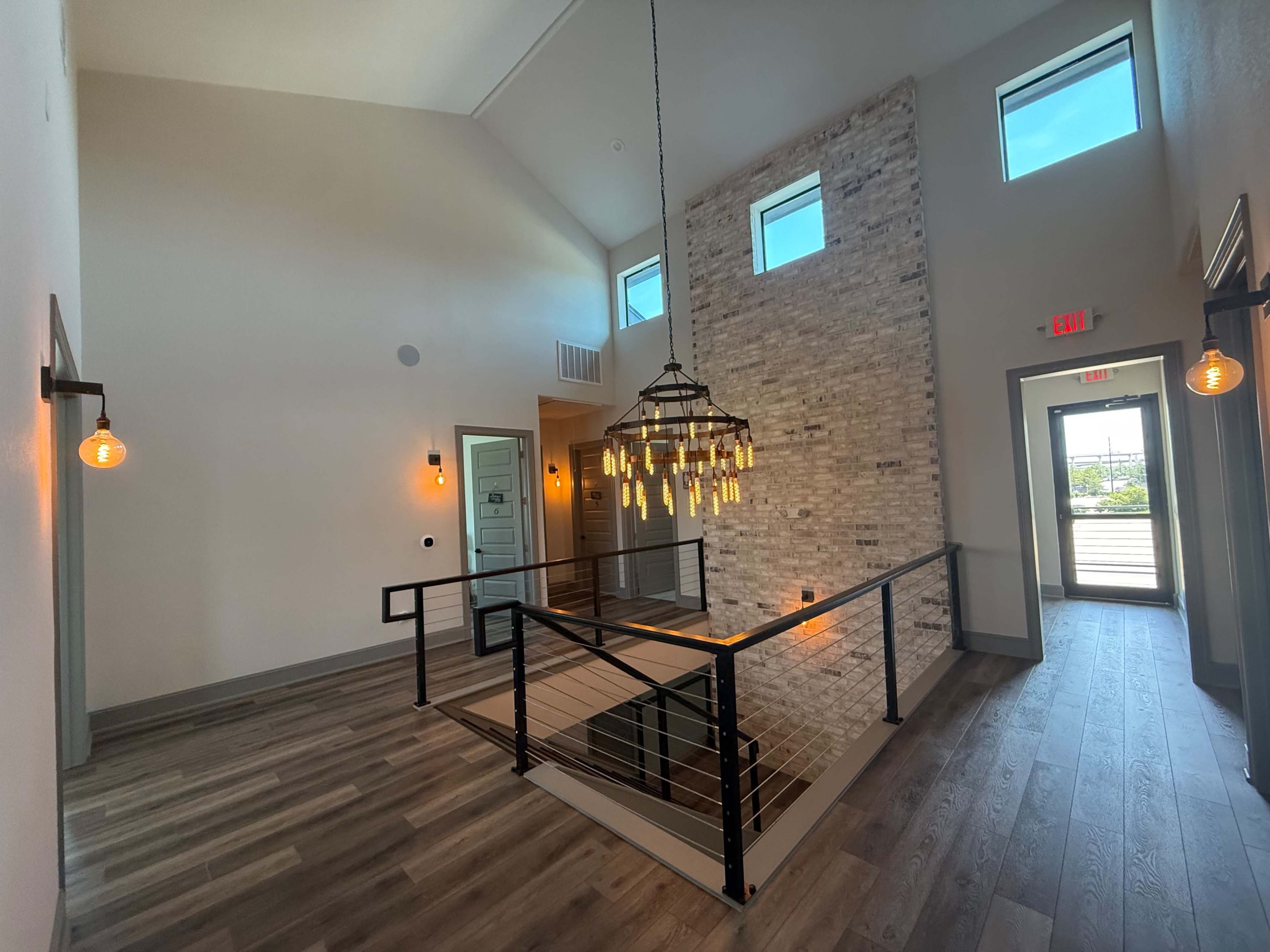 The image shows a modern two-story hallway with a wooden floor, a large hanging chandelier, and a brick accent wall, illuminated by natural light from windows.