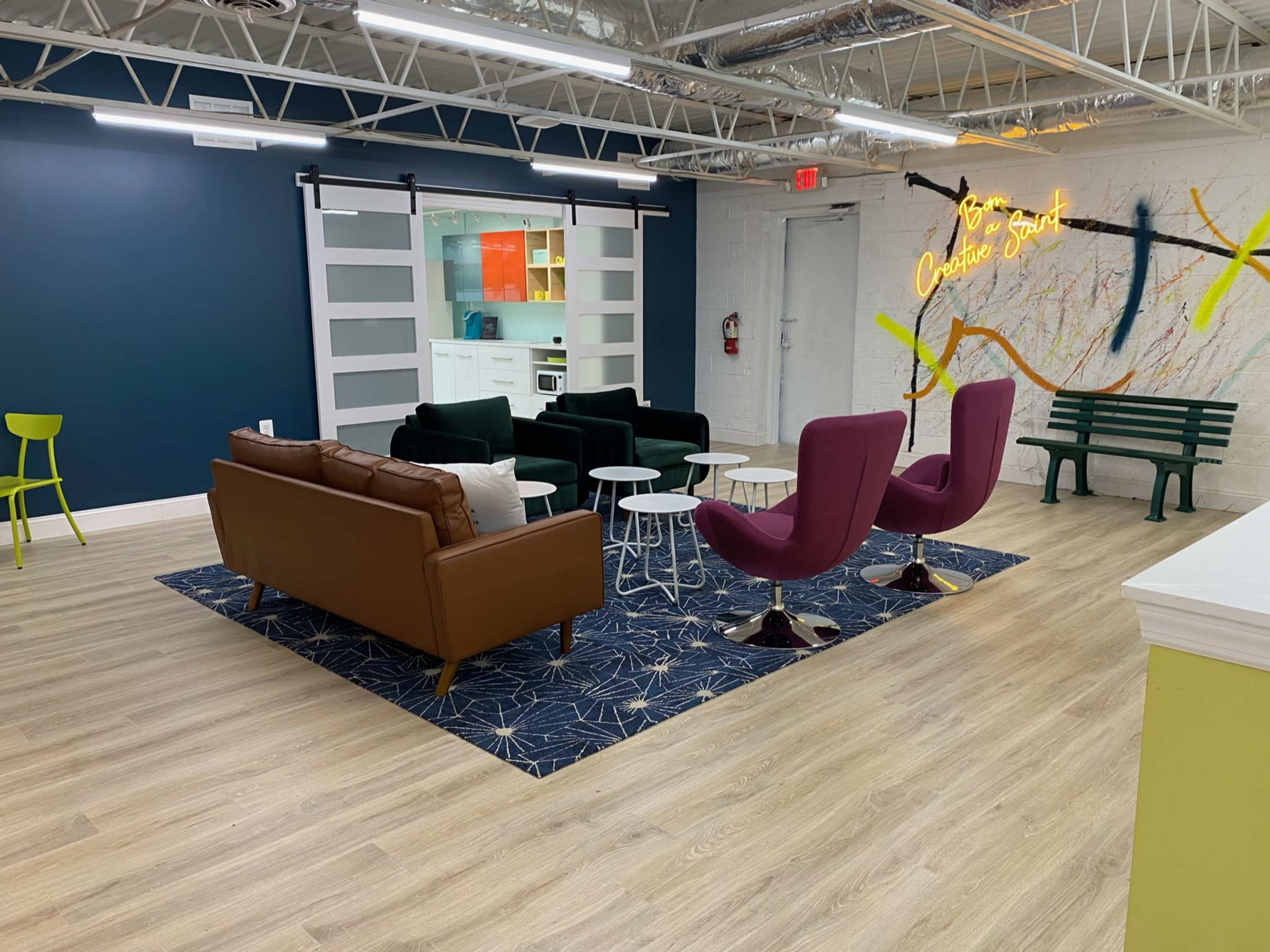 A modern lounge area with a mix of seating options, including a brown leather couch, purple armchairs, and a green bench, situated on a patterned rug in a well-lit space.
