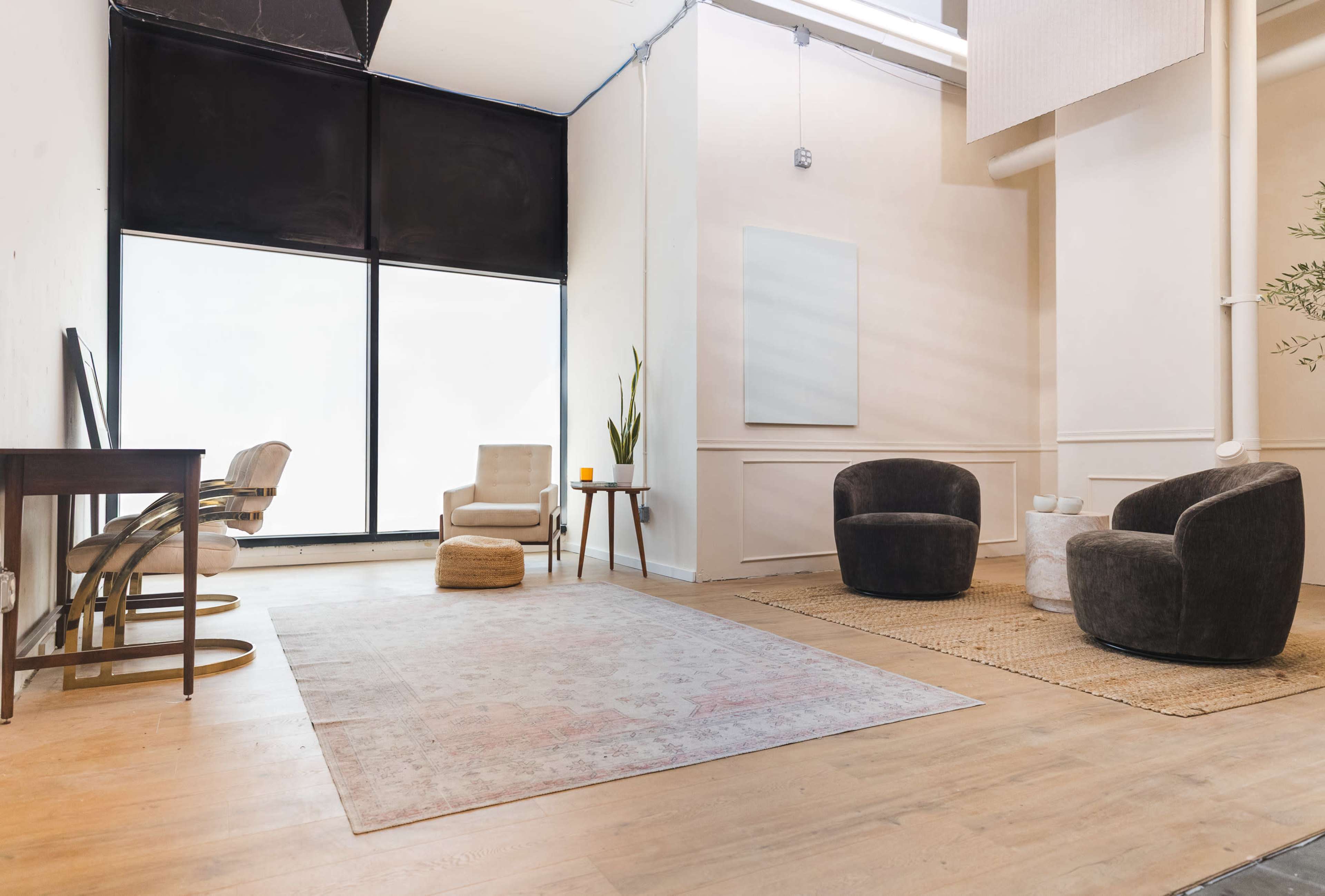 The image shows a minimalist interior space featuring two upholstered chairs, a small ottoman, a wooden table, and a large area rug on a wooden floor.