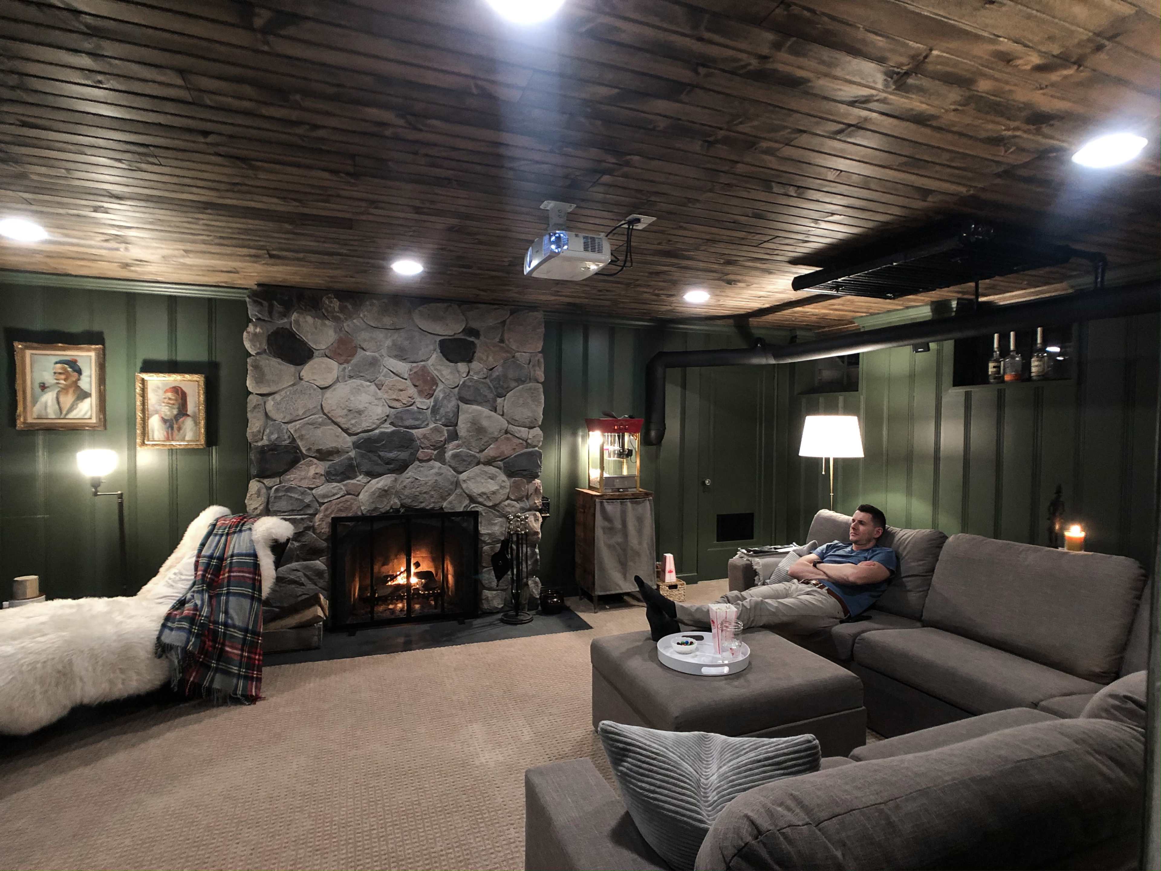 A cozy basement room features a stone fireplace, a projector on the ceiling, and a man sitting on a gray sectional couch amidst warm lighting and decorative elements.