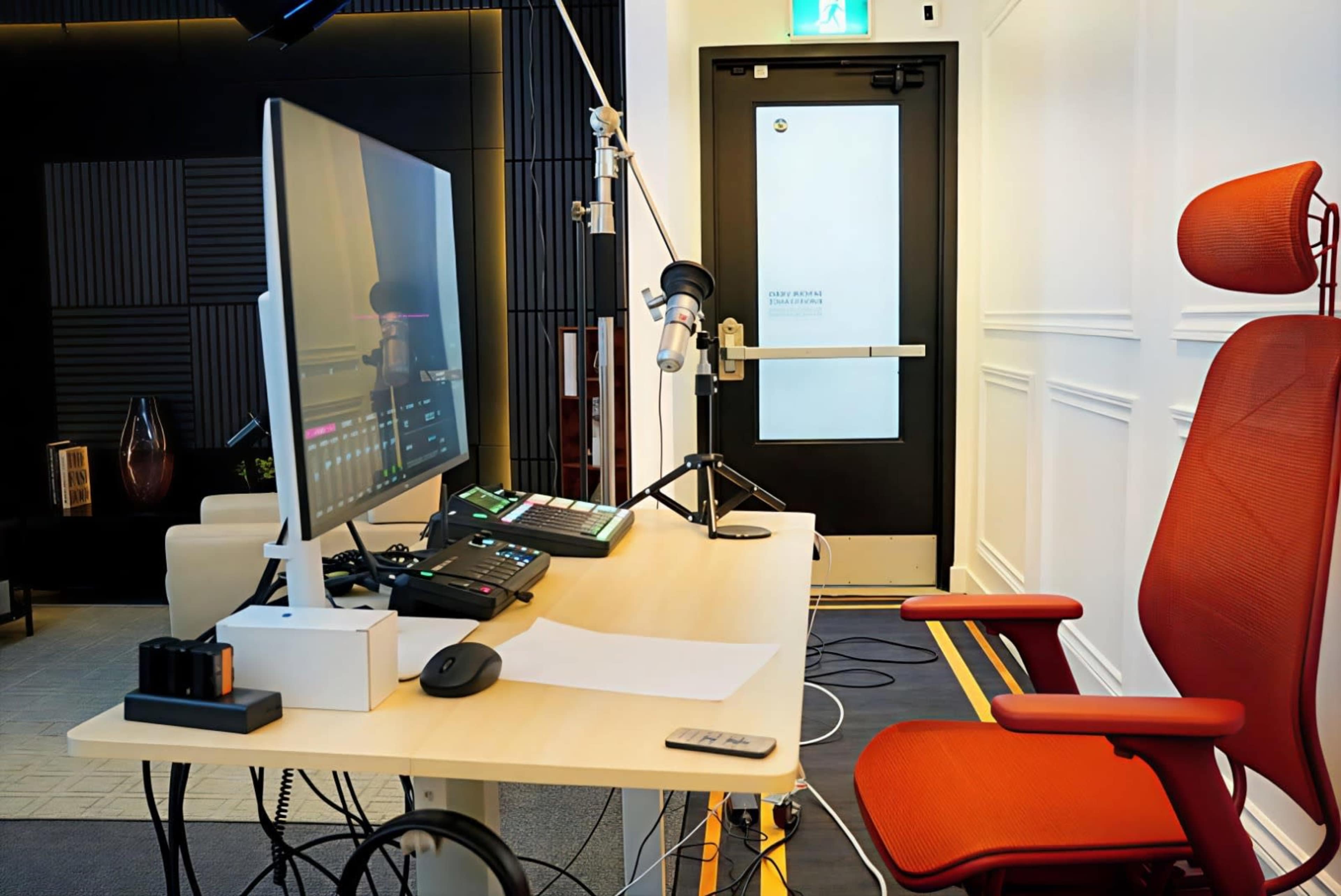 The image shows a modern workstation with a computer, microphone, and an ergonomic chair, positioned near a door in a well-lit room.