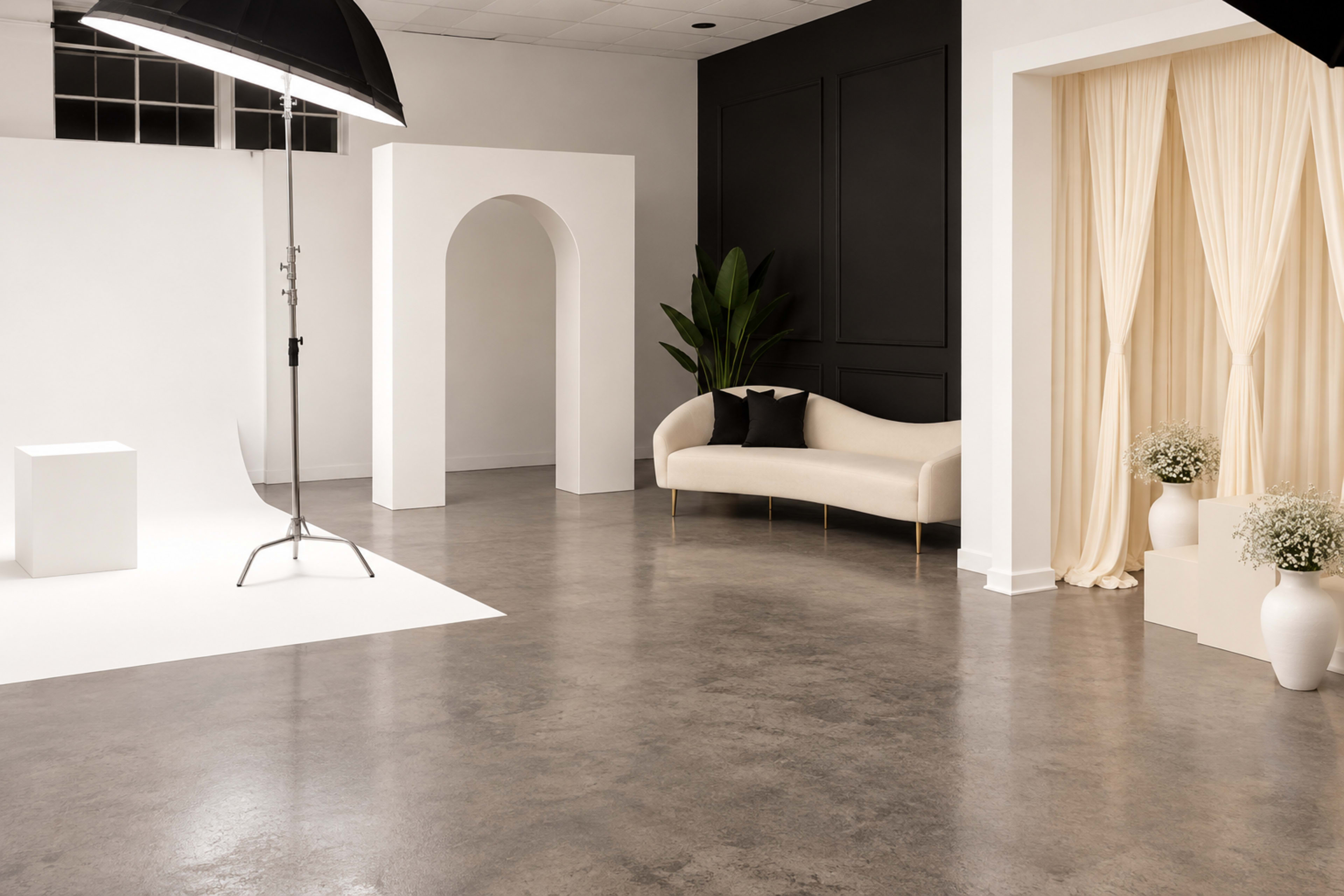 The image shows a modern studio interior featuring a neutral color palette, a curved white sofa, an archway, a backdrop for photography, and decorative elements like potted plants and vases.