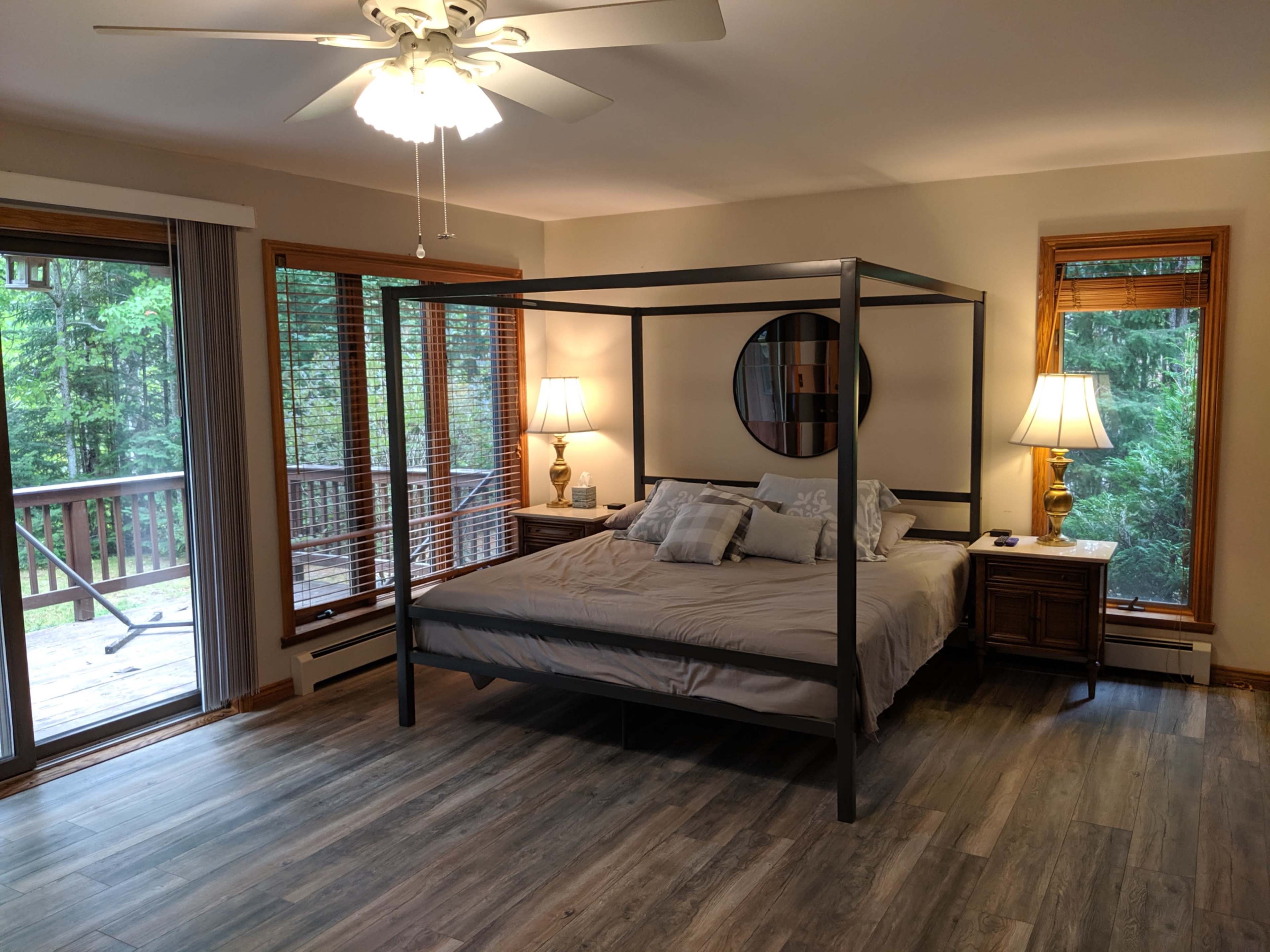 A spacious bedroom features a four-poster bed, two bedside tables with lamps, and large windows overlooking a wooded area.