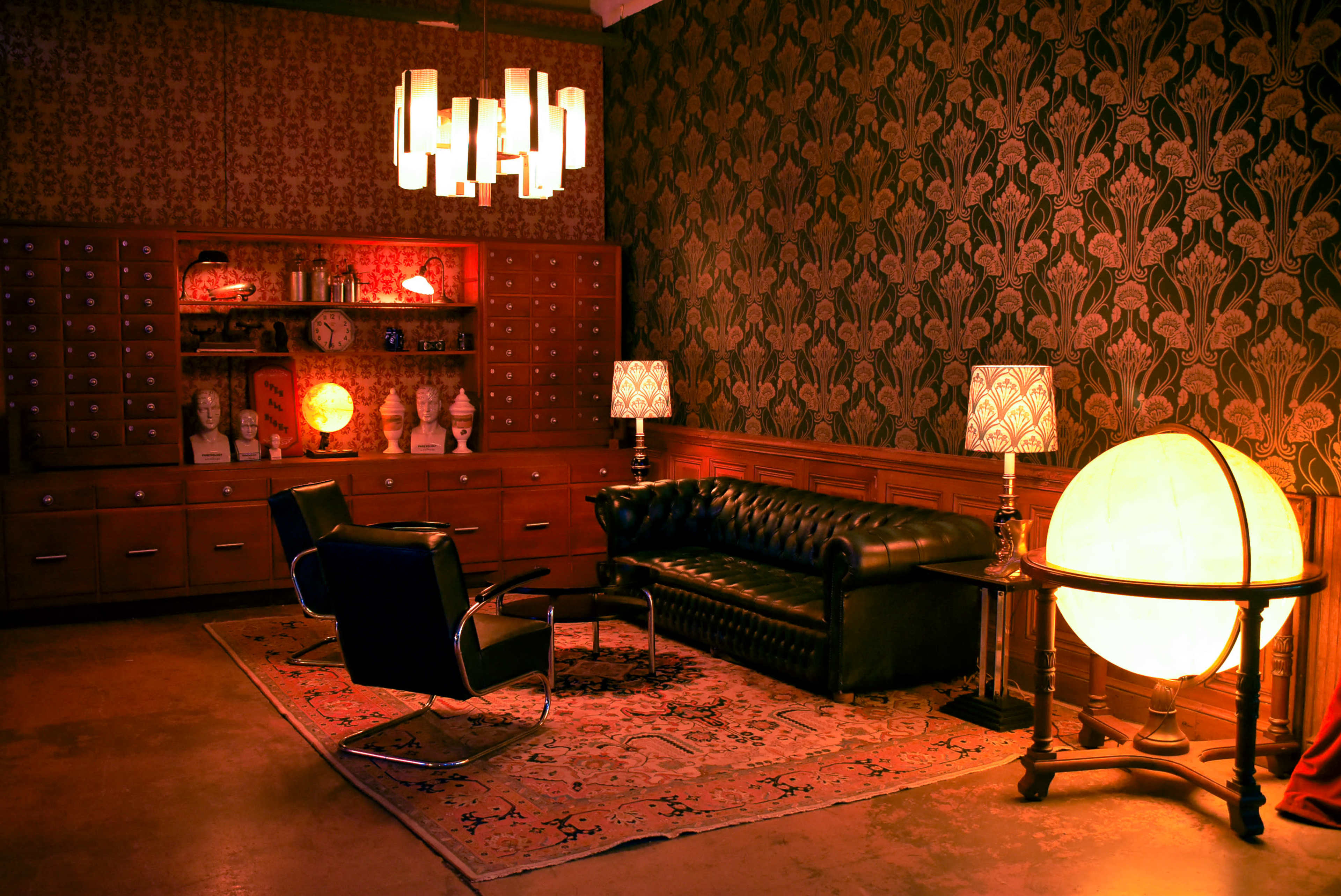 A stylish room with a black leather sofa, two modern chairs, a large globe lamp, and patterned wallpaper, illuminated by pendant lights and table lamps.