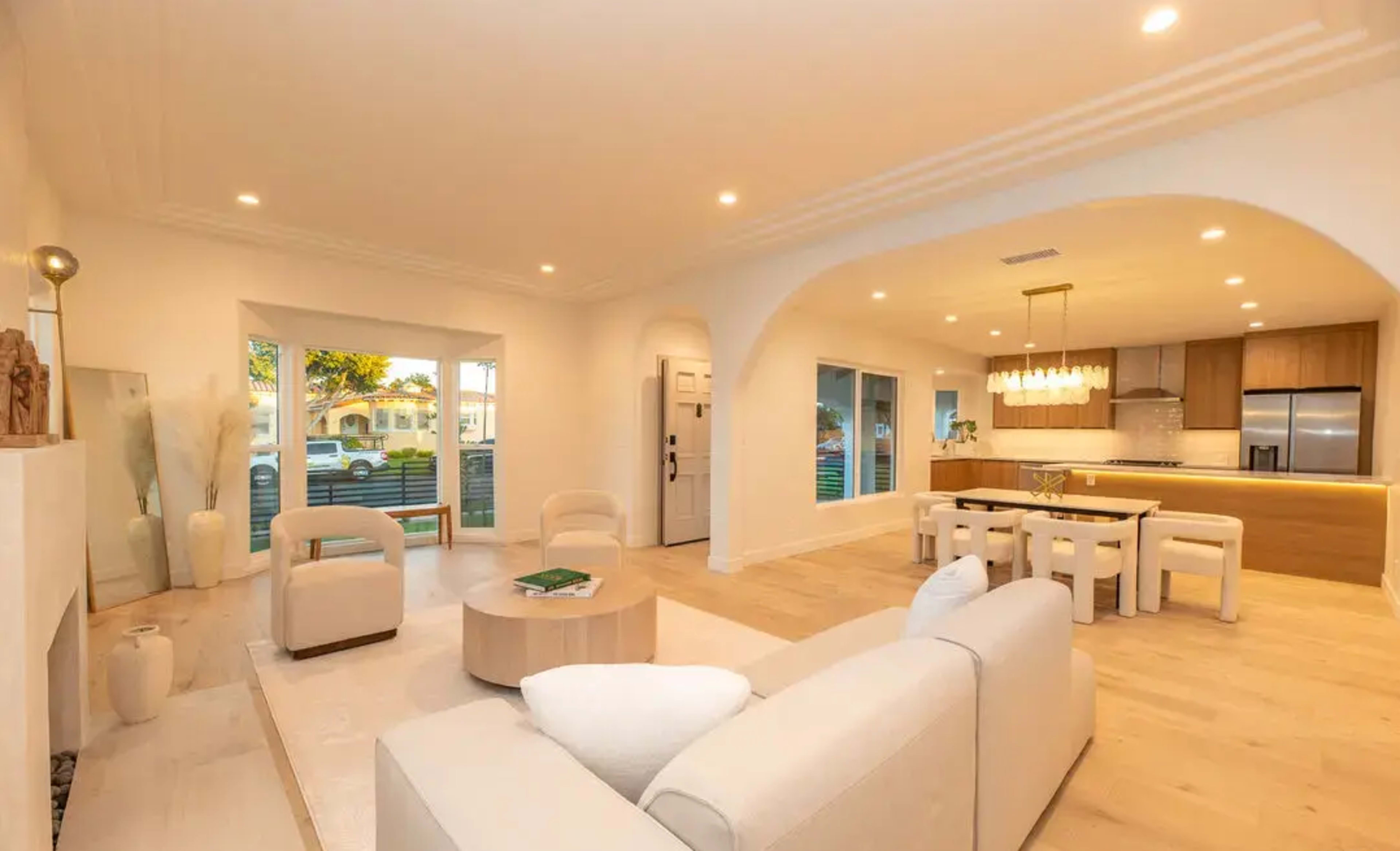 The image shows a bright, modern living area featuring a white sofa, a circular coffee table, and a dining space with a wooden table and chairs.