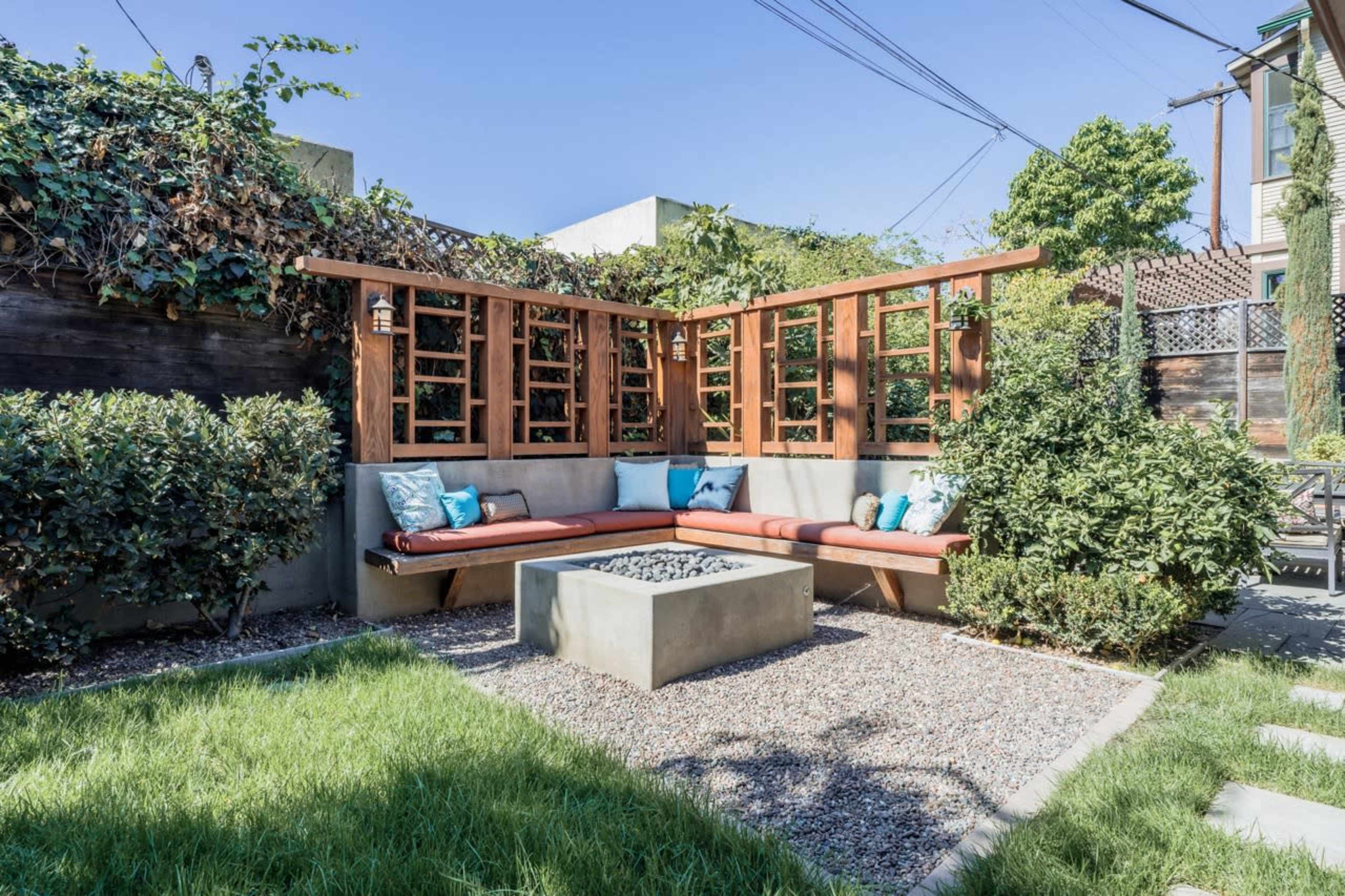 A modern outdoor seating area with a rectangular concrete fire pit, surrounded by greenery and a wooden trellis.