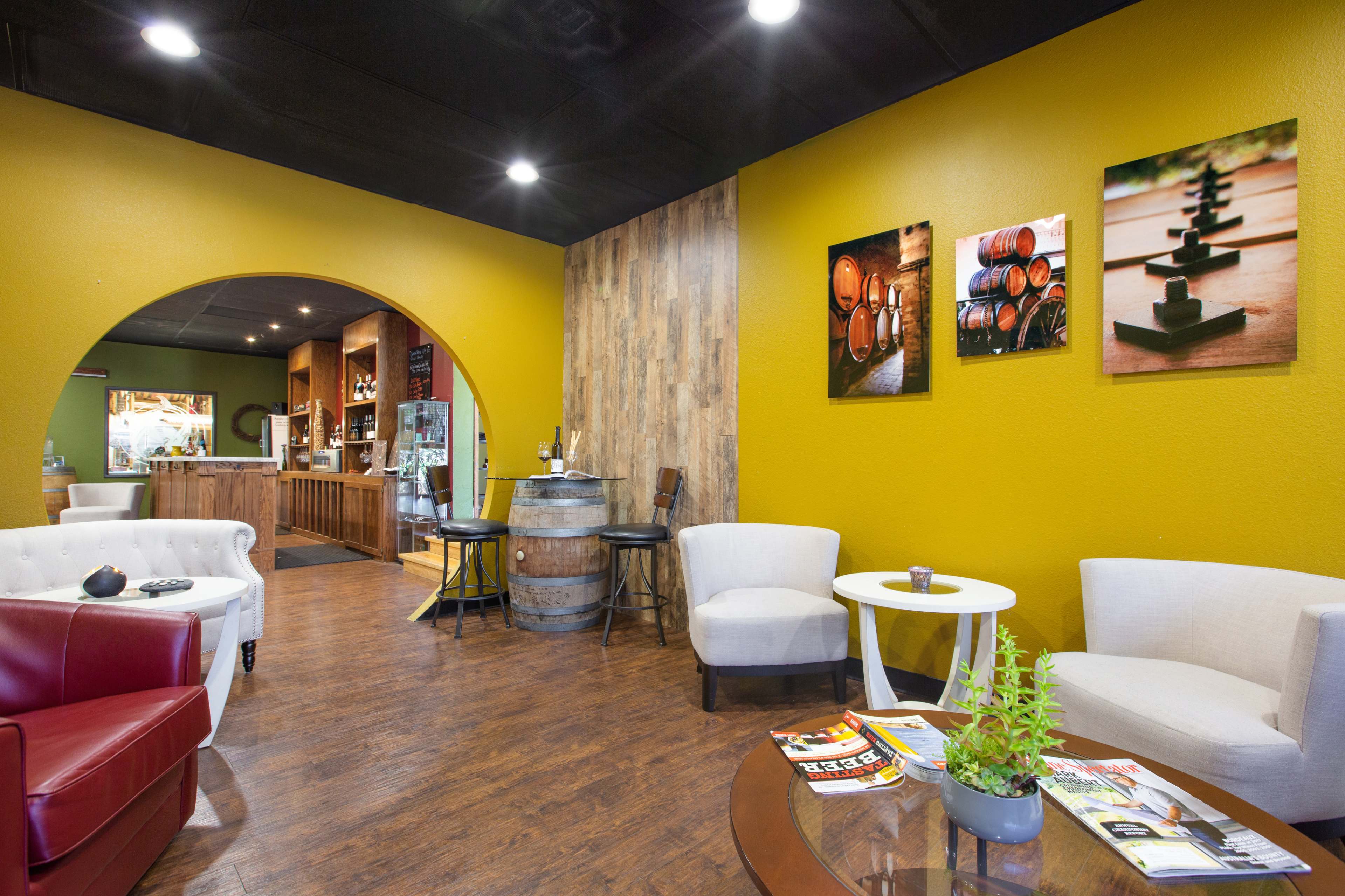 A cozy lounge area with two white chairs, a red chair, a round table, and a bar in the background, featuring wood accents and framed photographs on the walls.