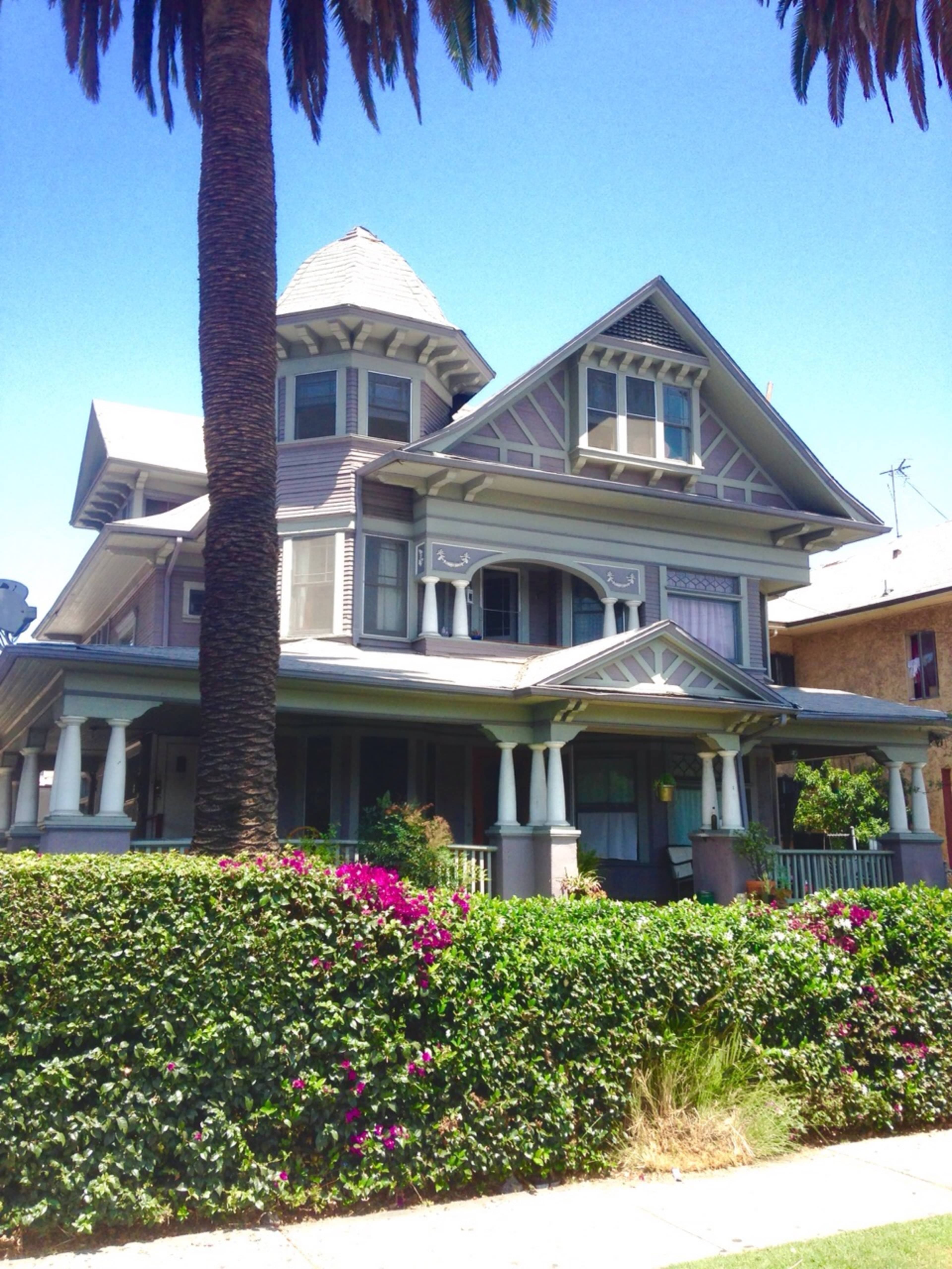 The image shows a large, multi-story Victorian-style house with a turret, surrounded by green shrubs and flowering plants, under a clear blue sky.