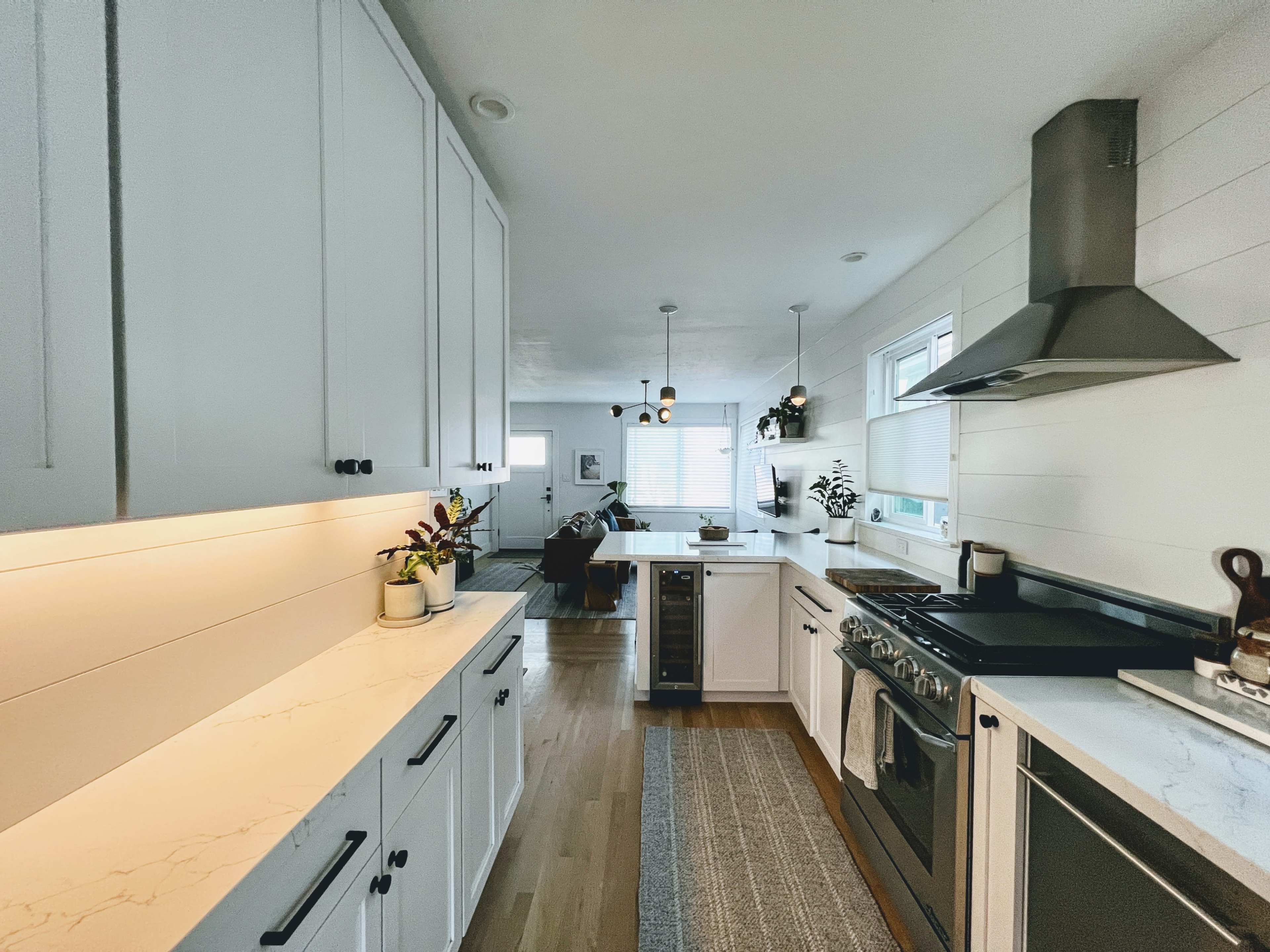 A modern kitchen features white cabinetry, marble countertops, stainless steel appliances, and pendant lighting, with a view into an adjoining living area.