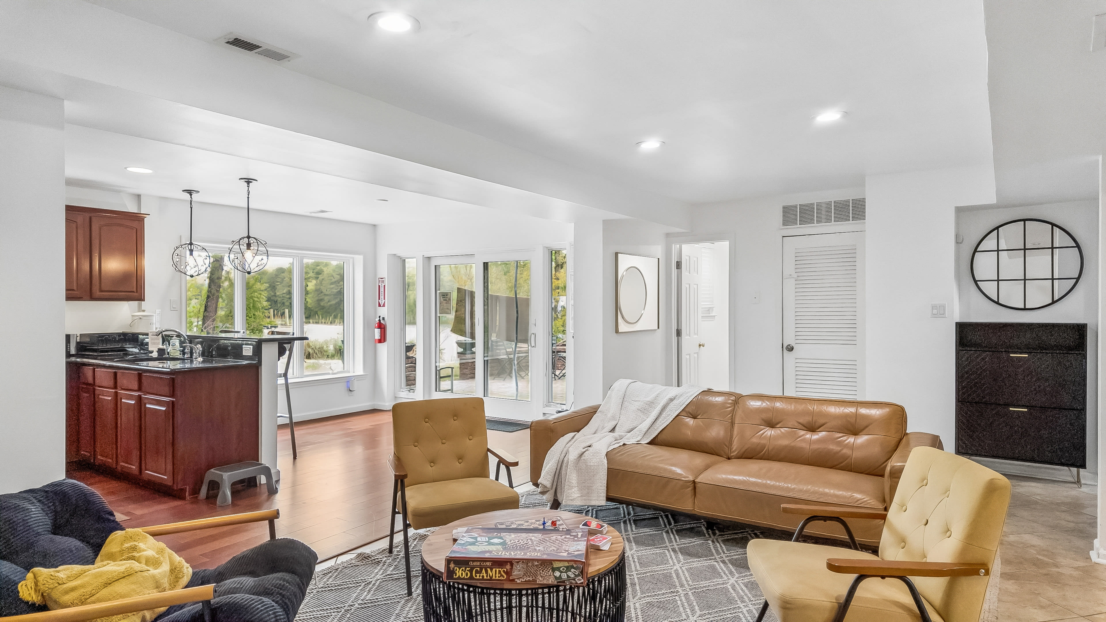 The image shows a spacious living area with a blend of seating options, a kitchen in the background, and large glass doors leading to an outdoor space.