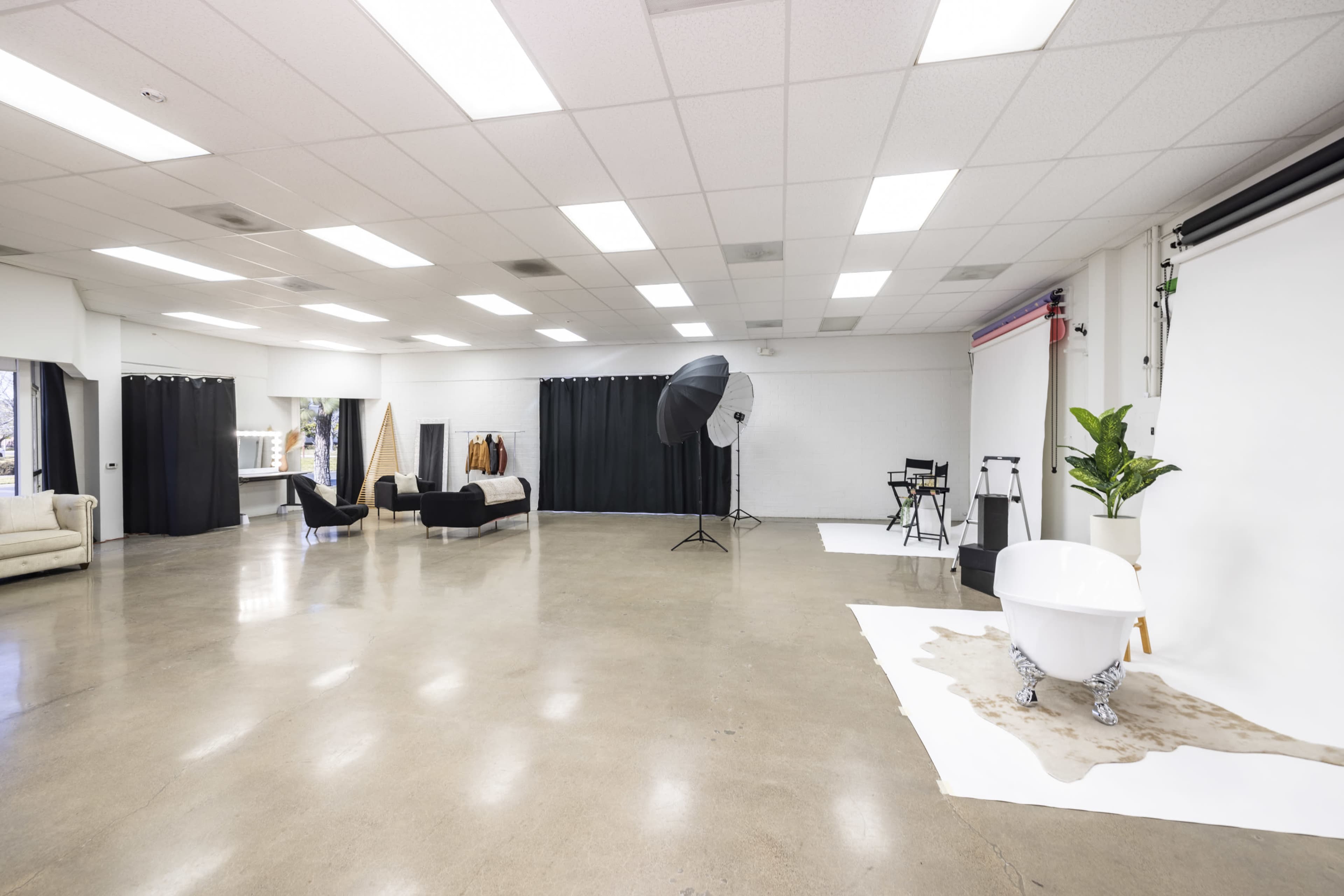 A spacious photography studio features a white backdrop, a large softbox light, and various props including a bathtub and clothing on hangers.