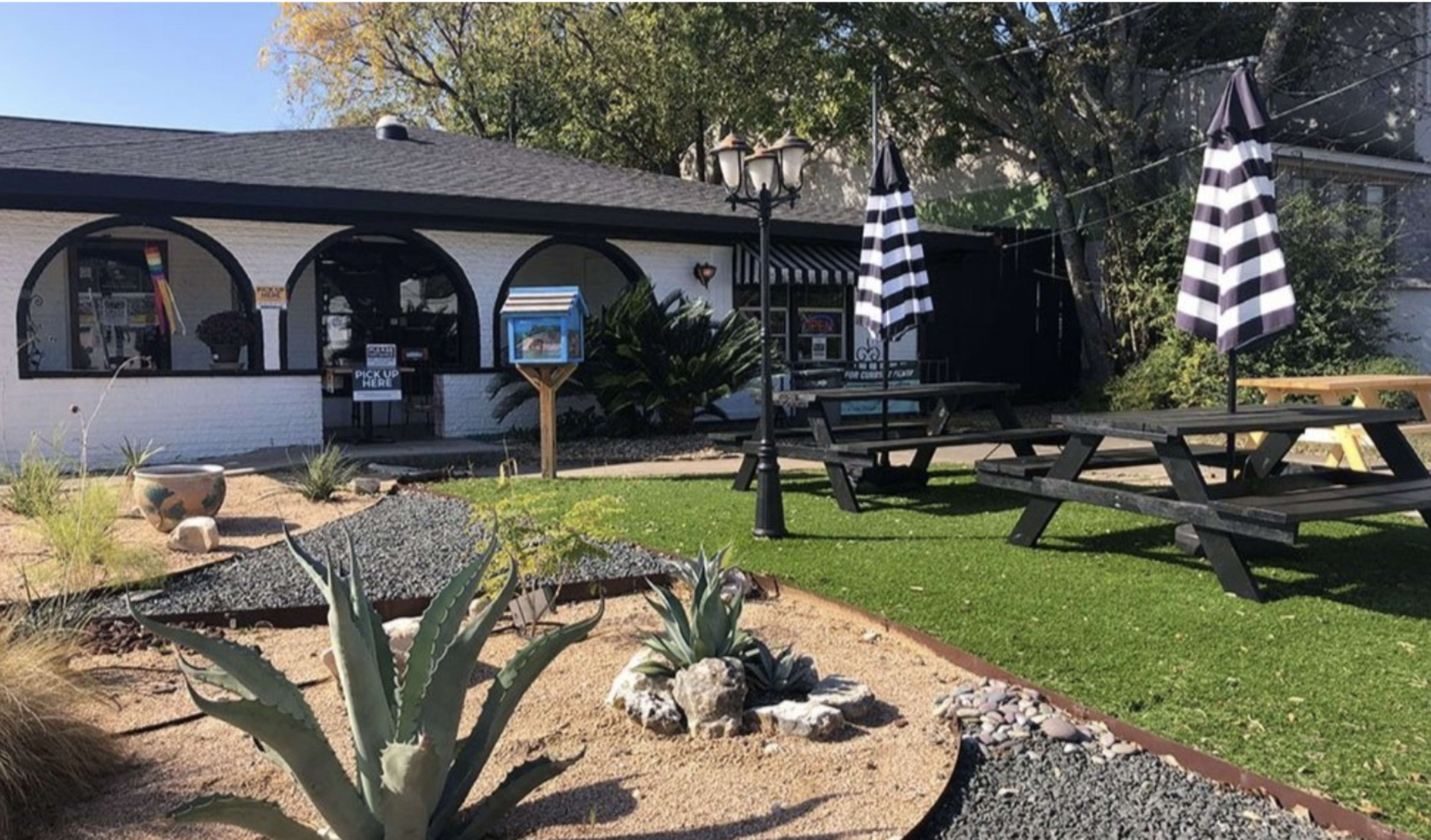The image shows a landscaped outdoor area with black and white striped umbrellas over picnic tables, bordered by gravel and plants in front of a white building.