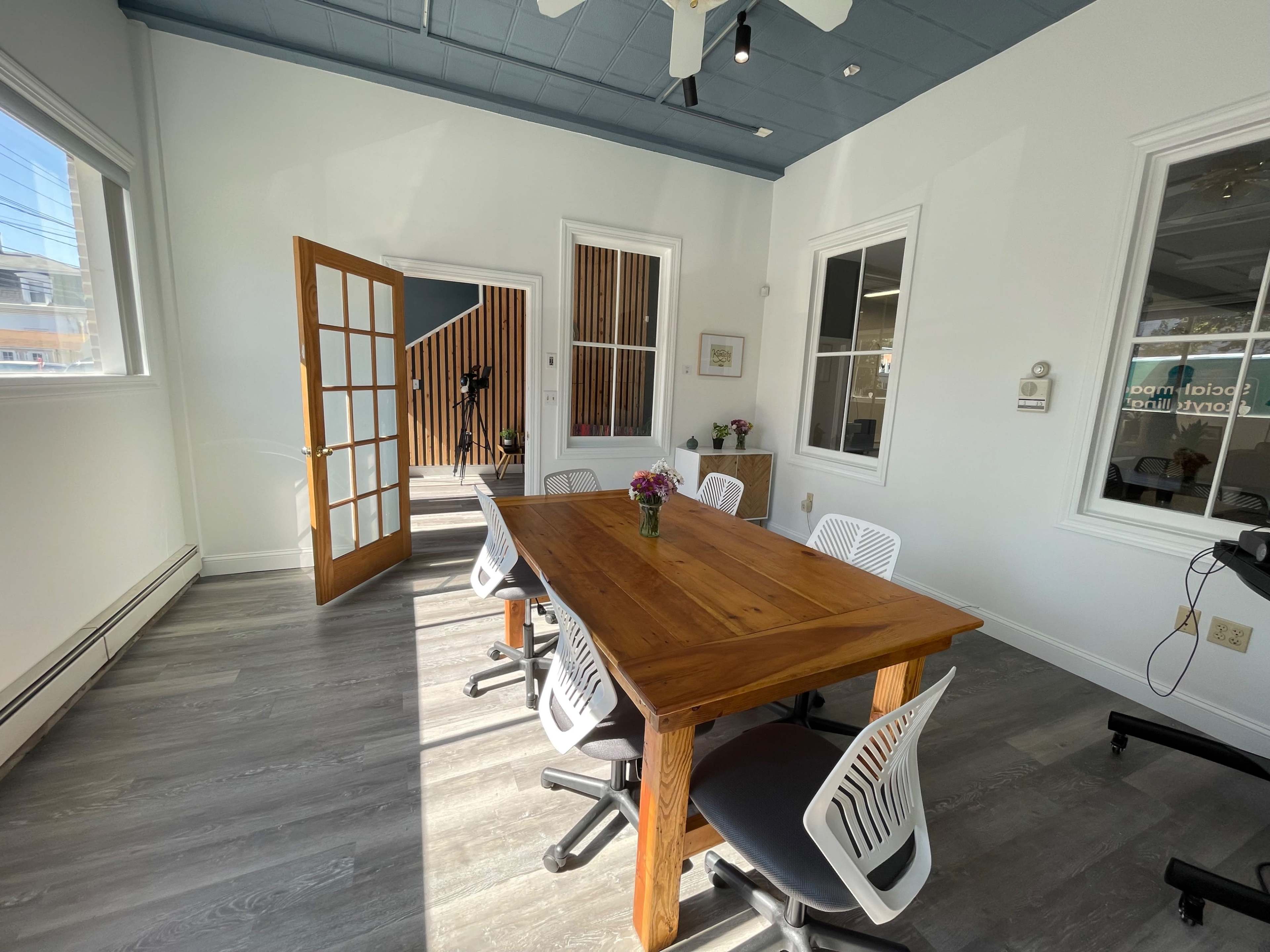 A wooden table surrounded by six chairs is positioned in a bright meeting room with large windows and an open door leading to a staircase.