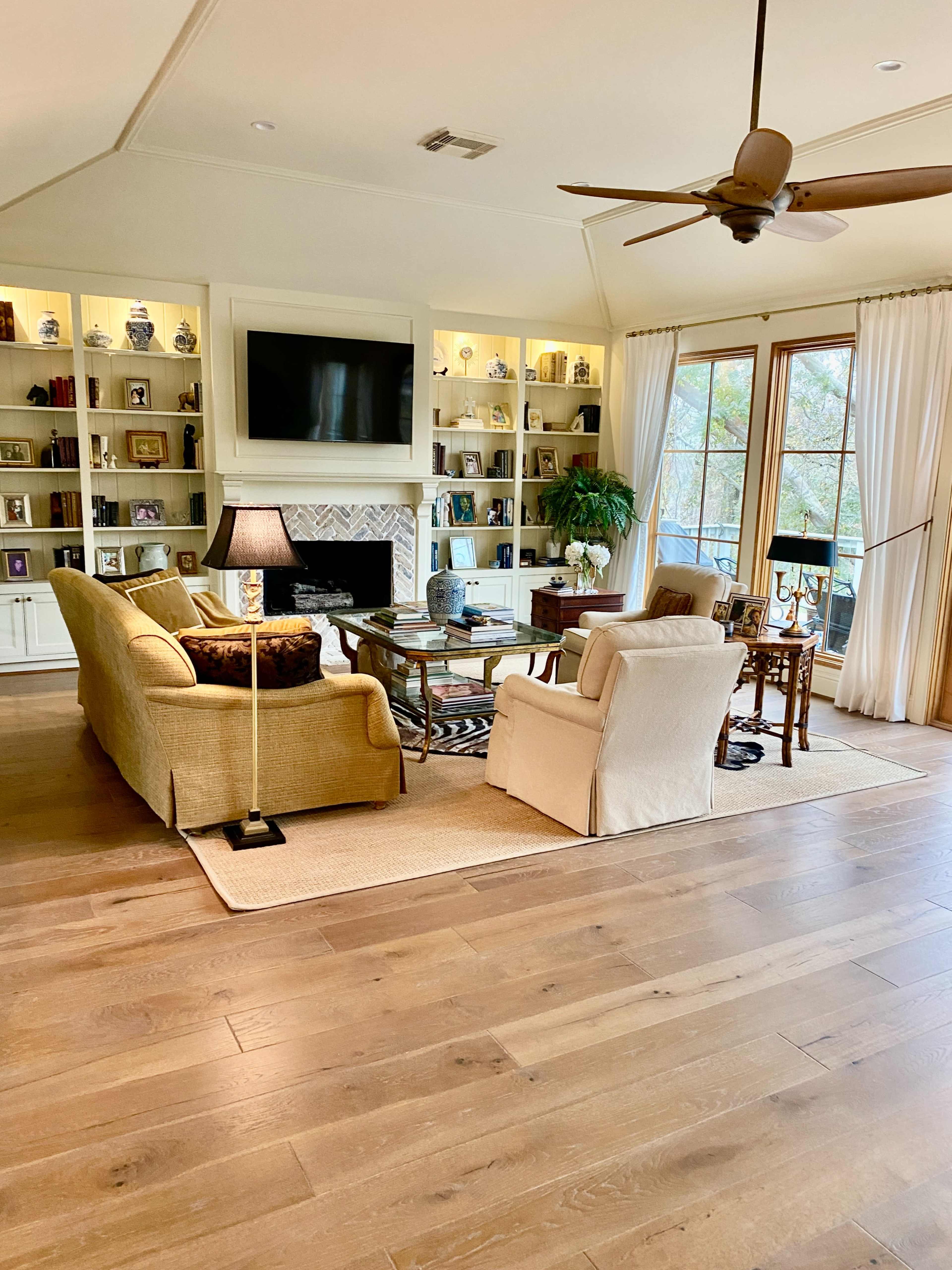 A spacious living room features a seating area with a sofa, armchairs, a coffee table, and built-in shelves filled with books and decor, all set against large windows that provide natural light.