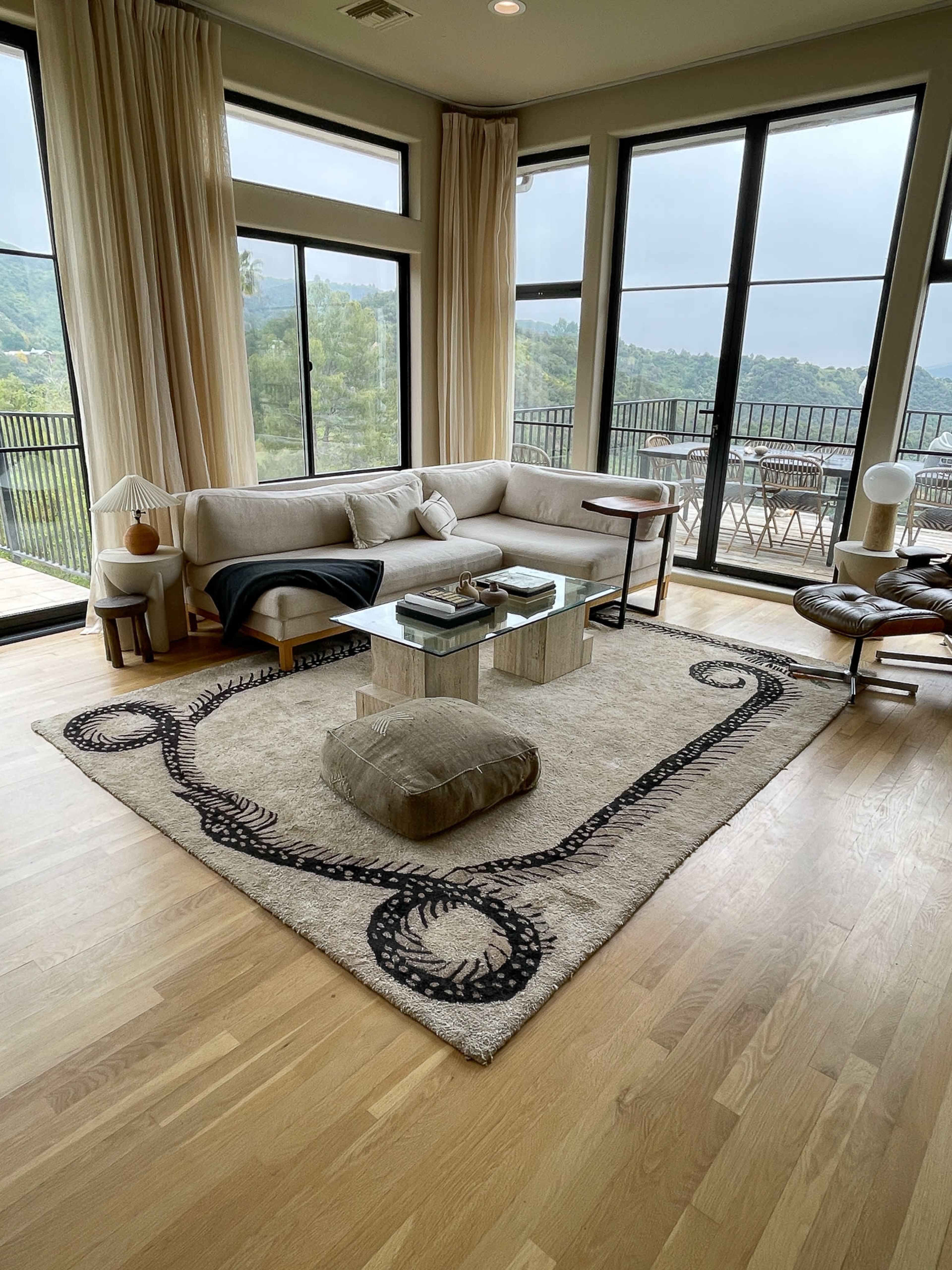 A modern living room with large windows, a soft beige sofa, a glass coffee table, a round floor cushion, and a patterned area rug.