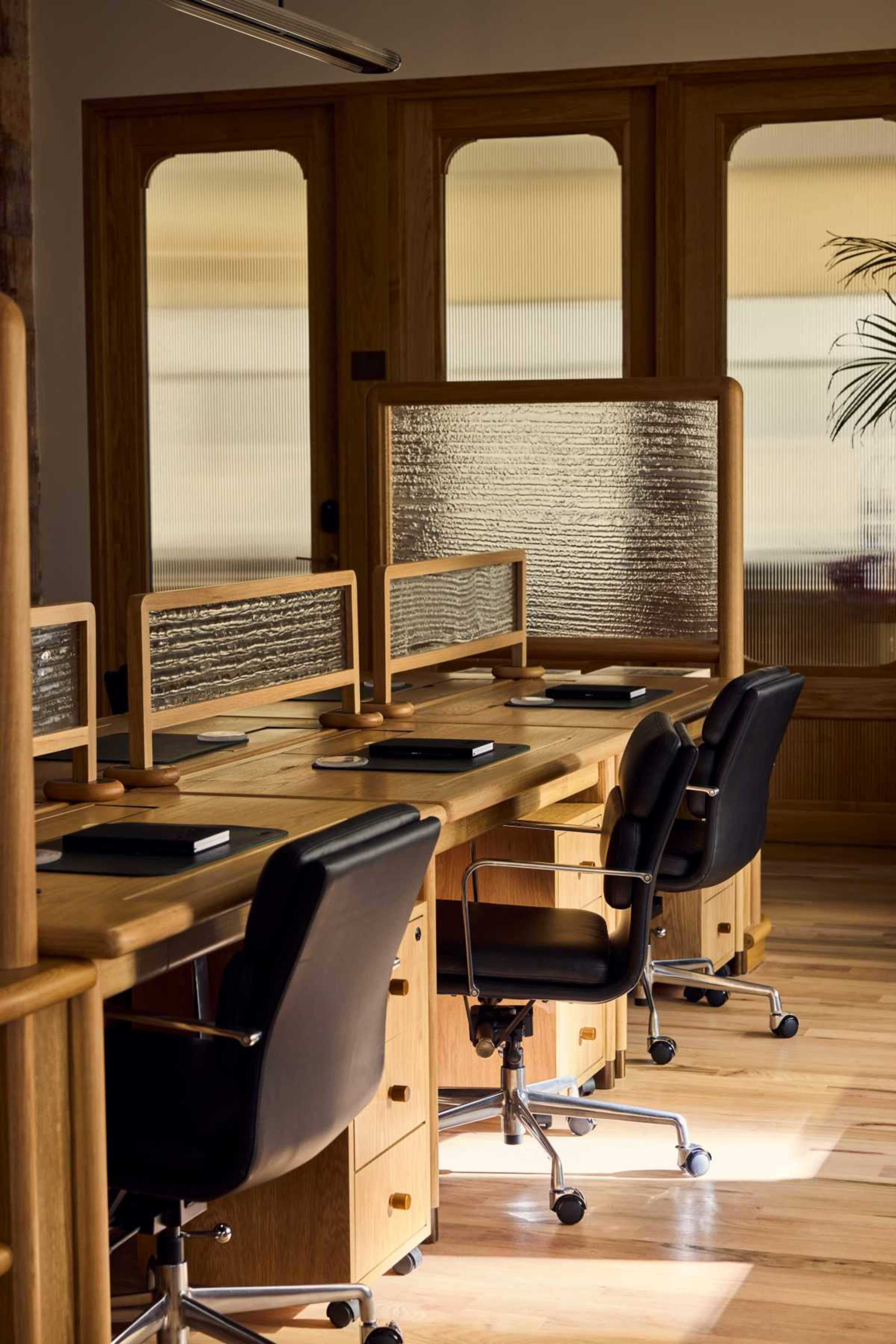 The image shows a stylish office workspace with several wooden desks equipped with black chairs and dividers, illuminated by natural light from large windows.