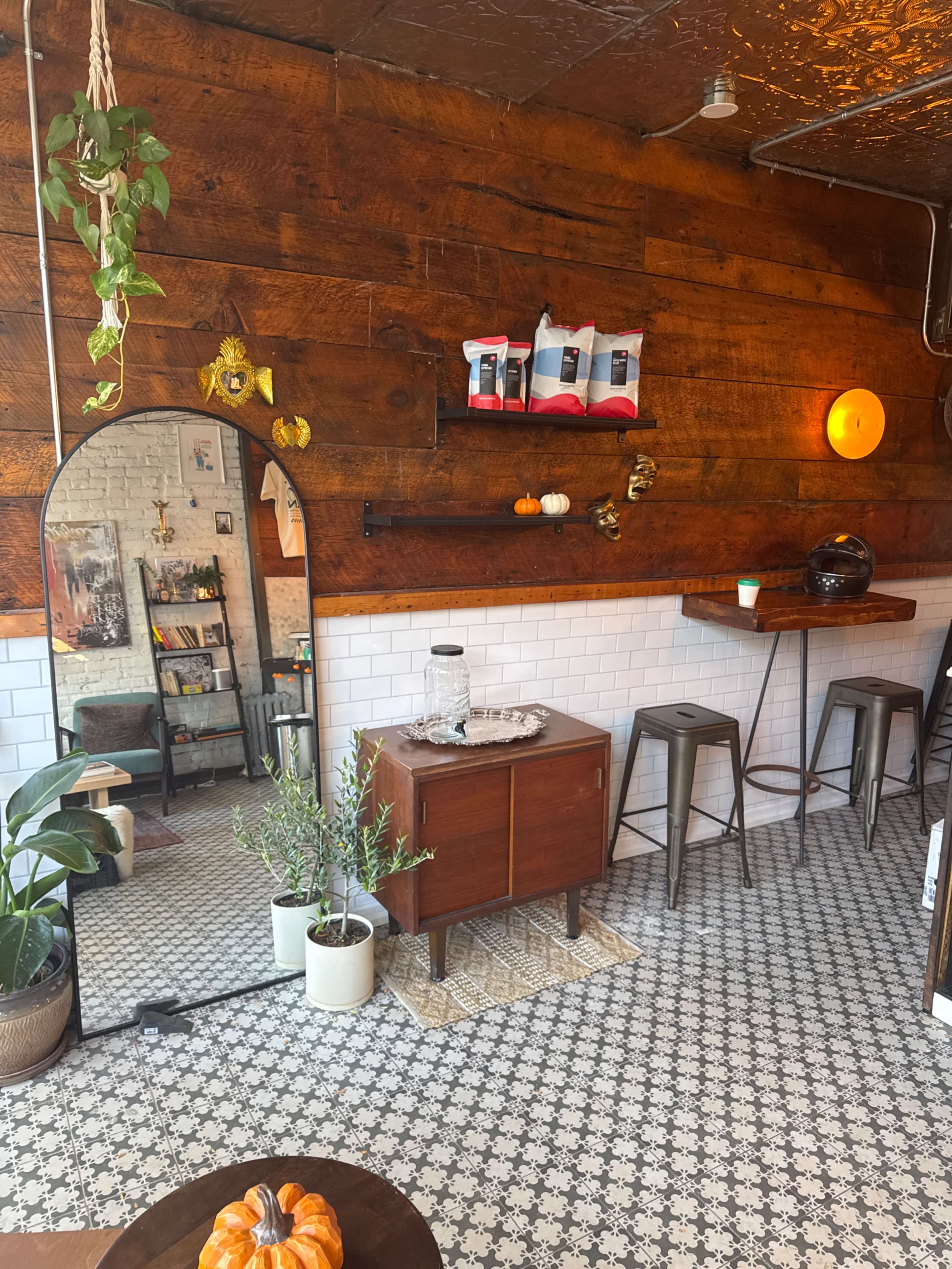 The image shows a cozy café interior with wooden walls, patterned tile flooring, and a mix of plants, furniture, and decorative items.