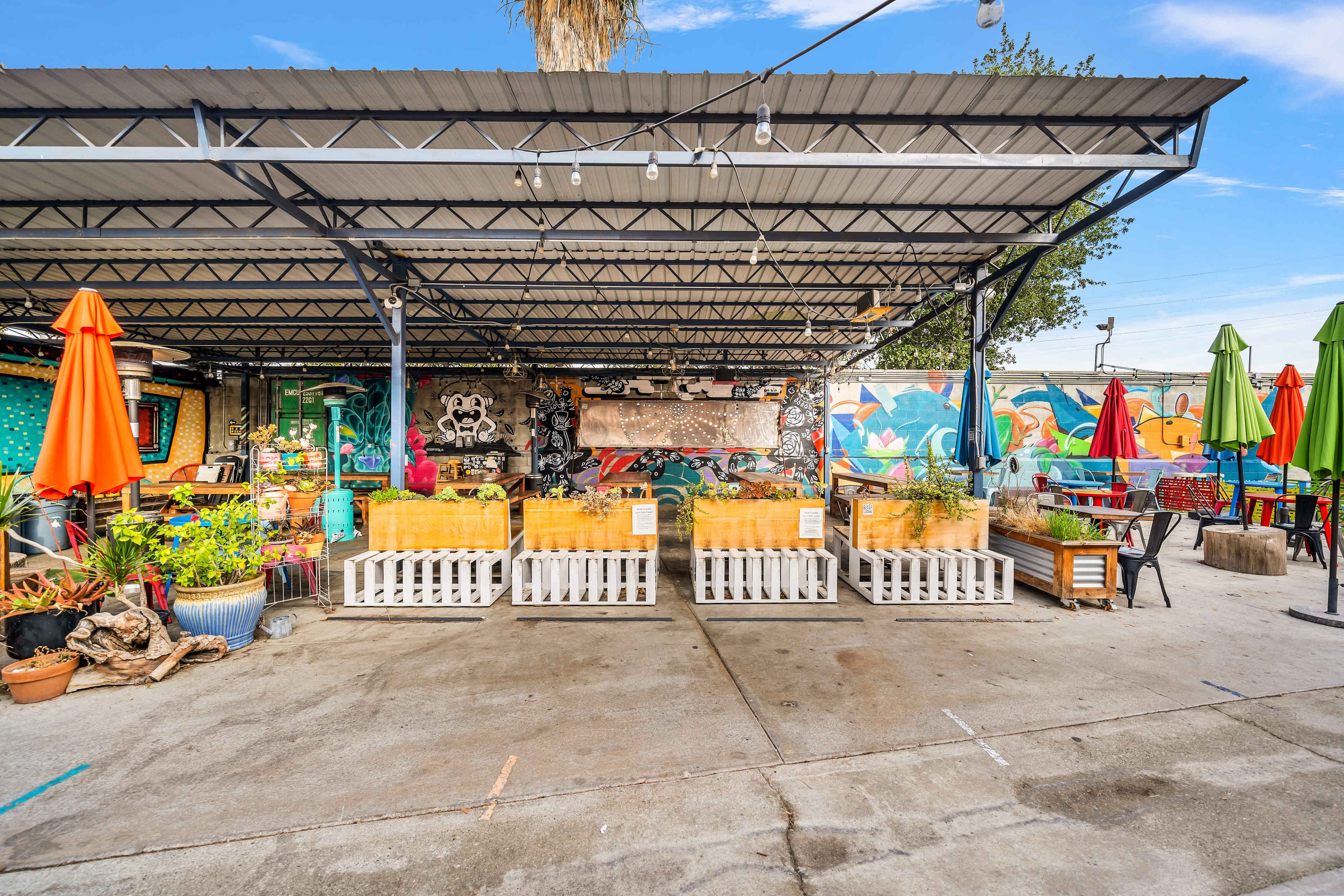 An outdoor seating area with colorful umbrellas, surrounded by vibrant murals and various potted plants.