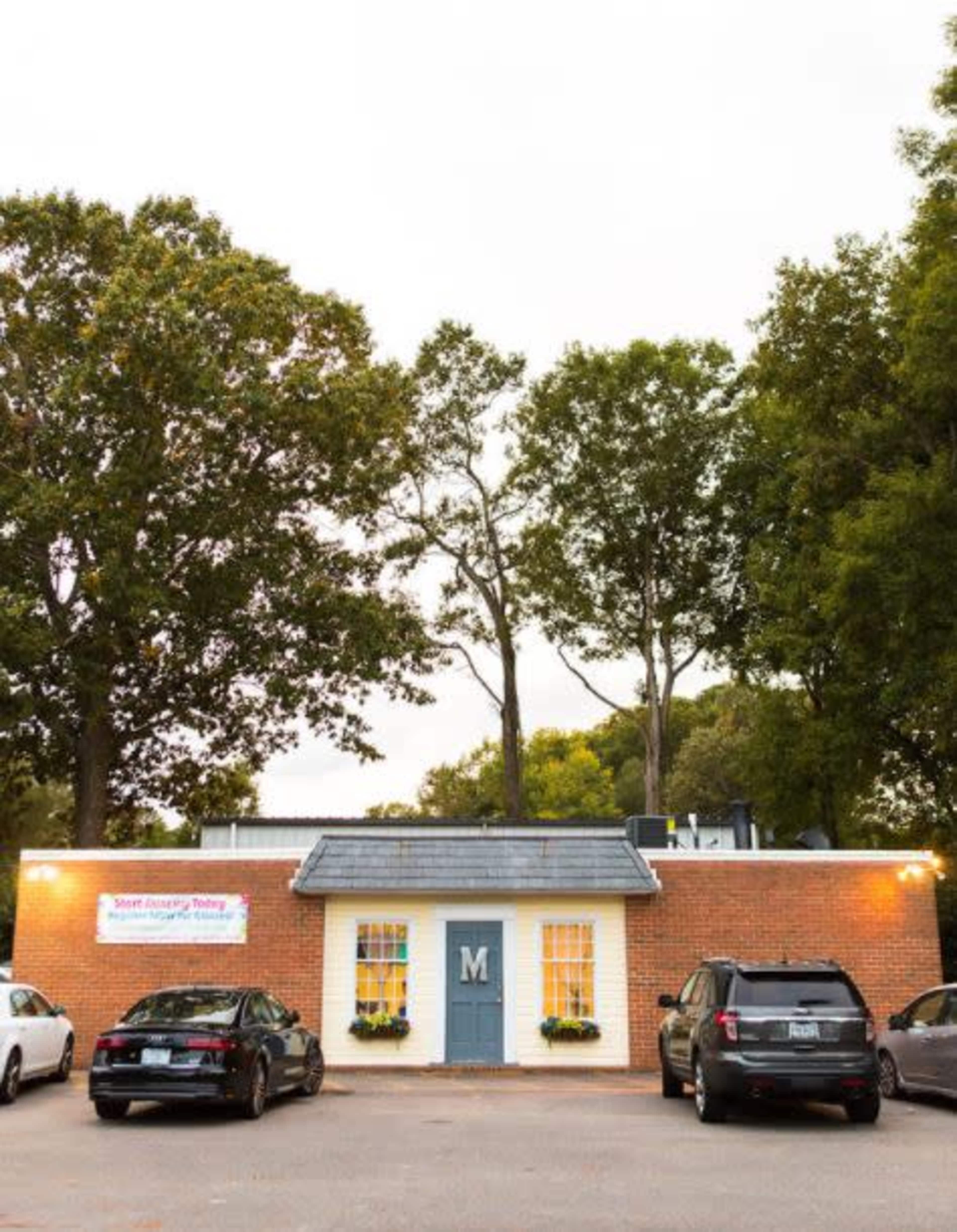 A brick building with a central door marked with the letter "M," surrounded by several parked cars and trees in the background.