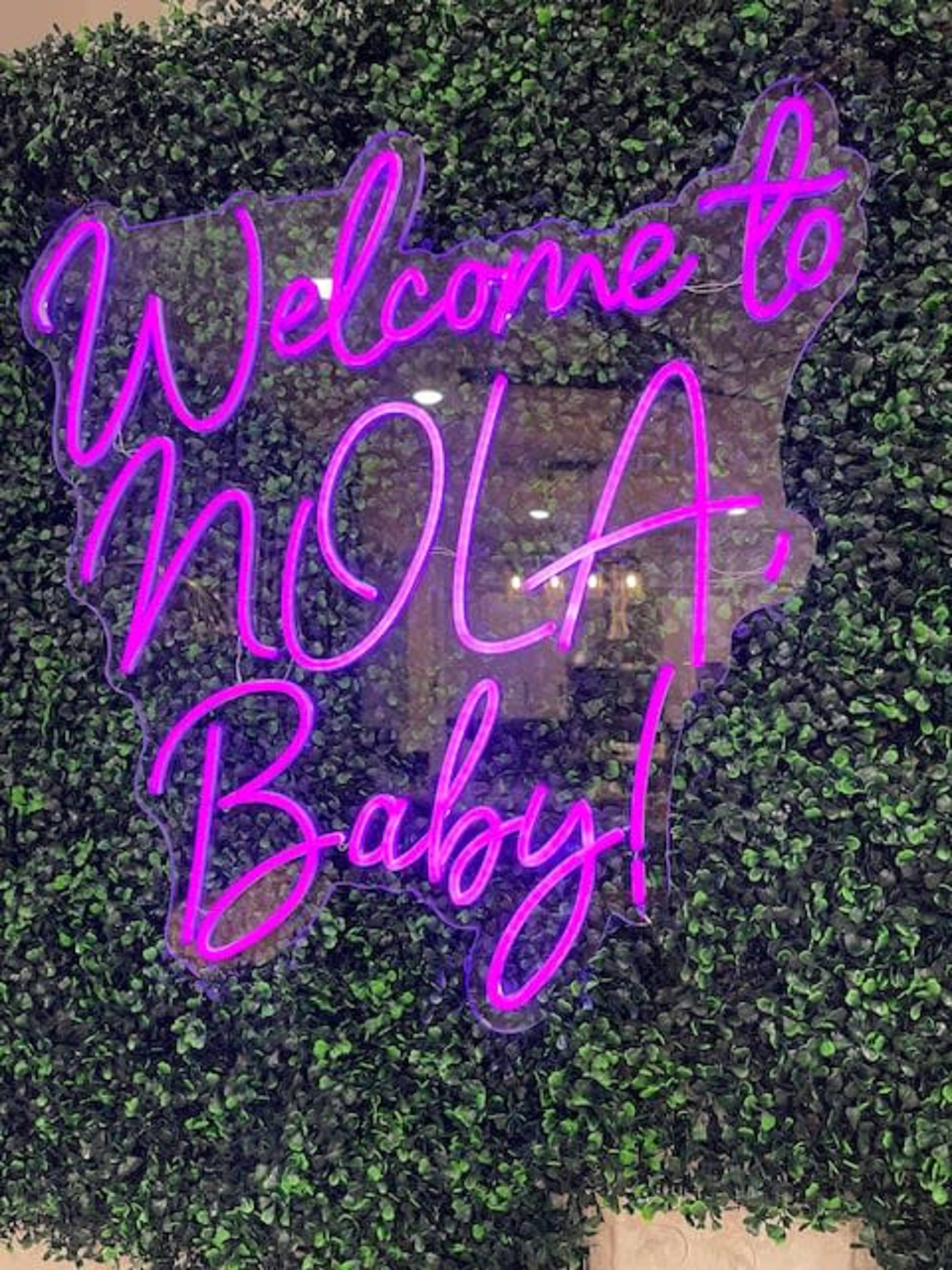 A neon sign reads "Welcome to NOLA Baby!" against a backdrop of green foliage.