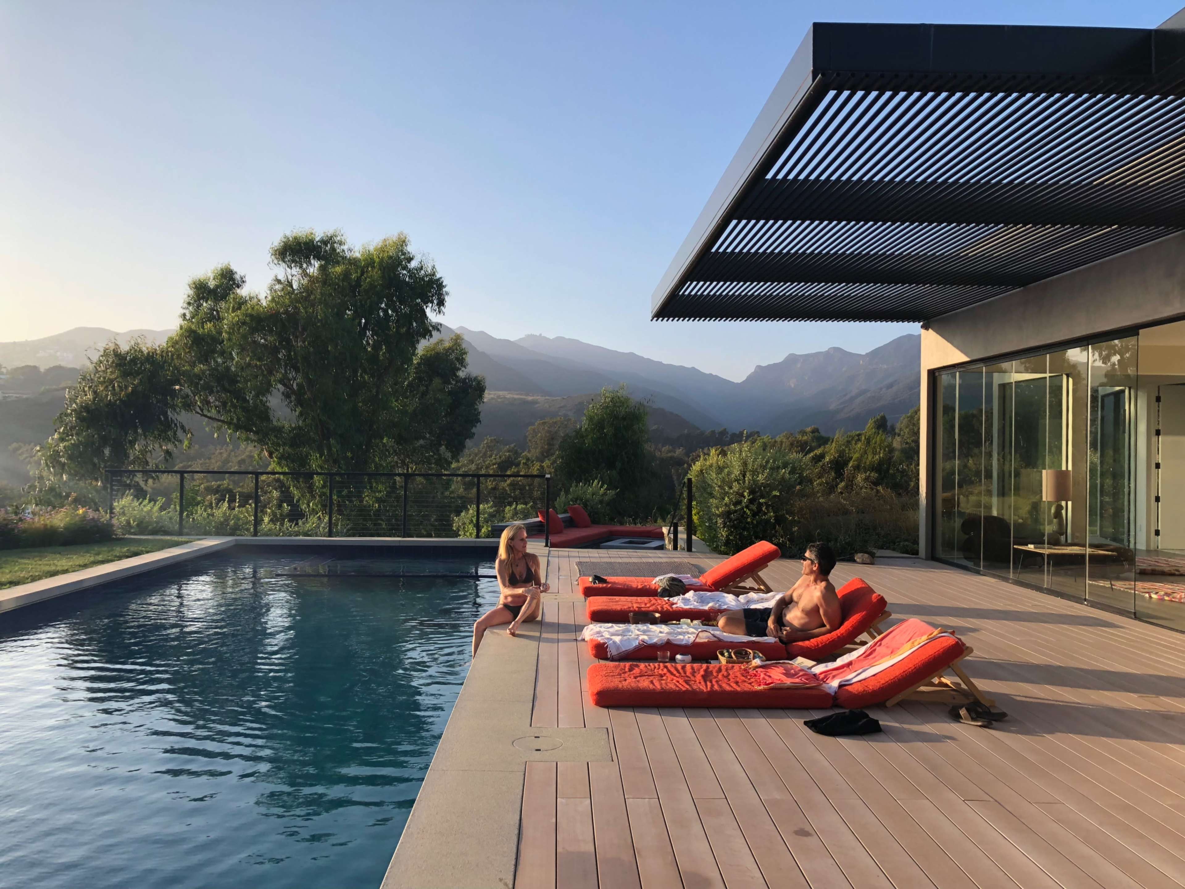 Two people relax on lounge chairs beside a swimming pool, with mountains visible in the background during sunset.