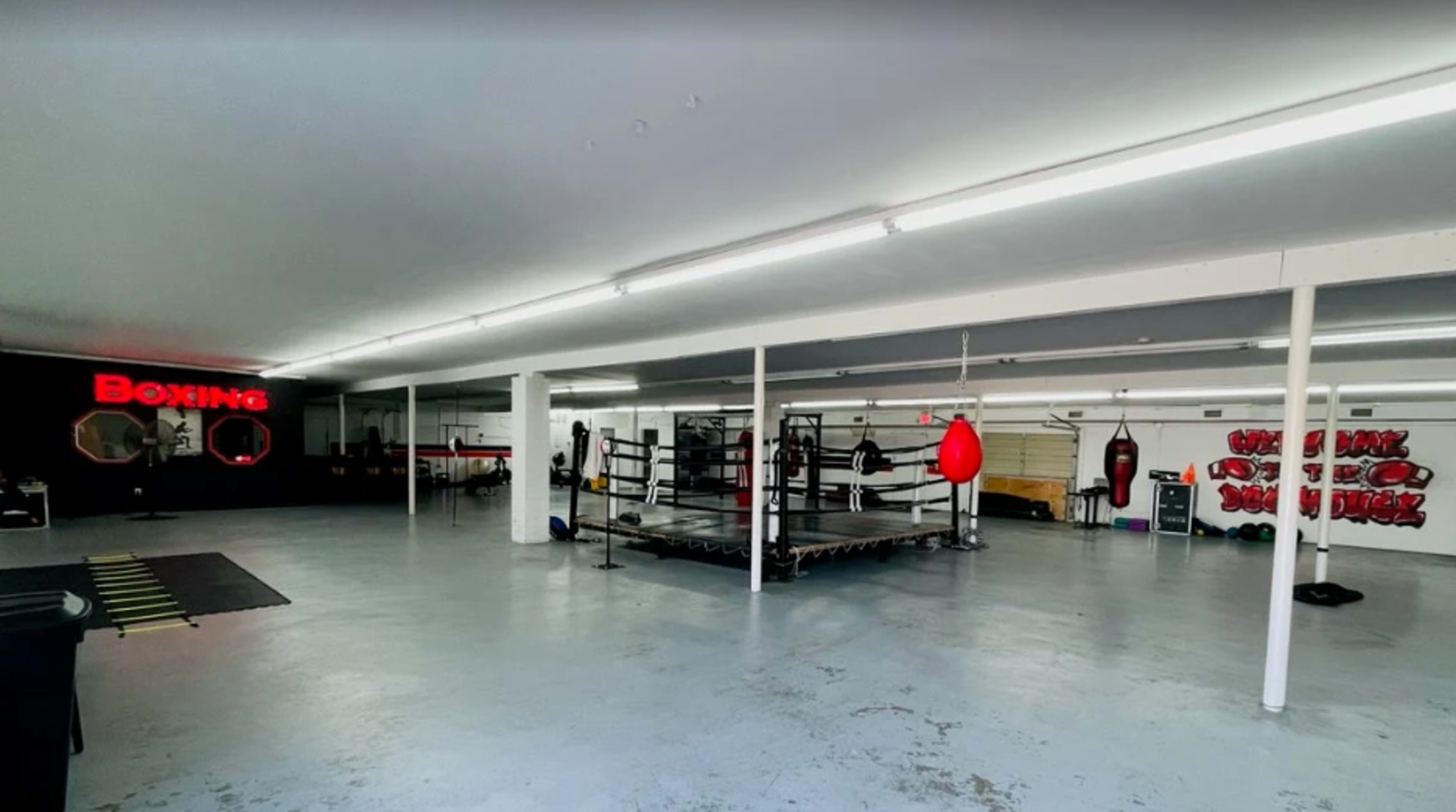 The image shows a spacious boxing gym equipped with a boxing ring, training bags, and workout equipment, featuring a minimalist design and bright lighting.
