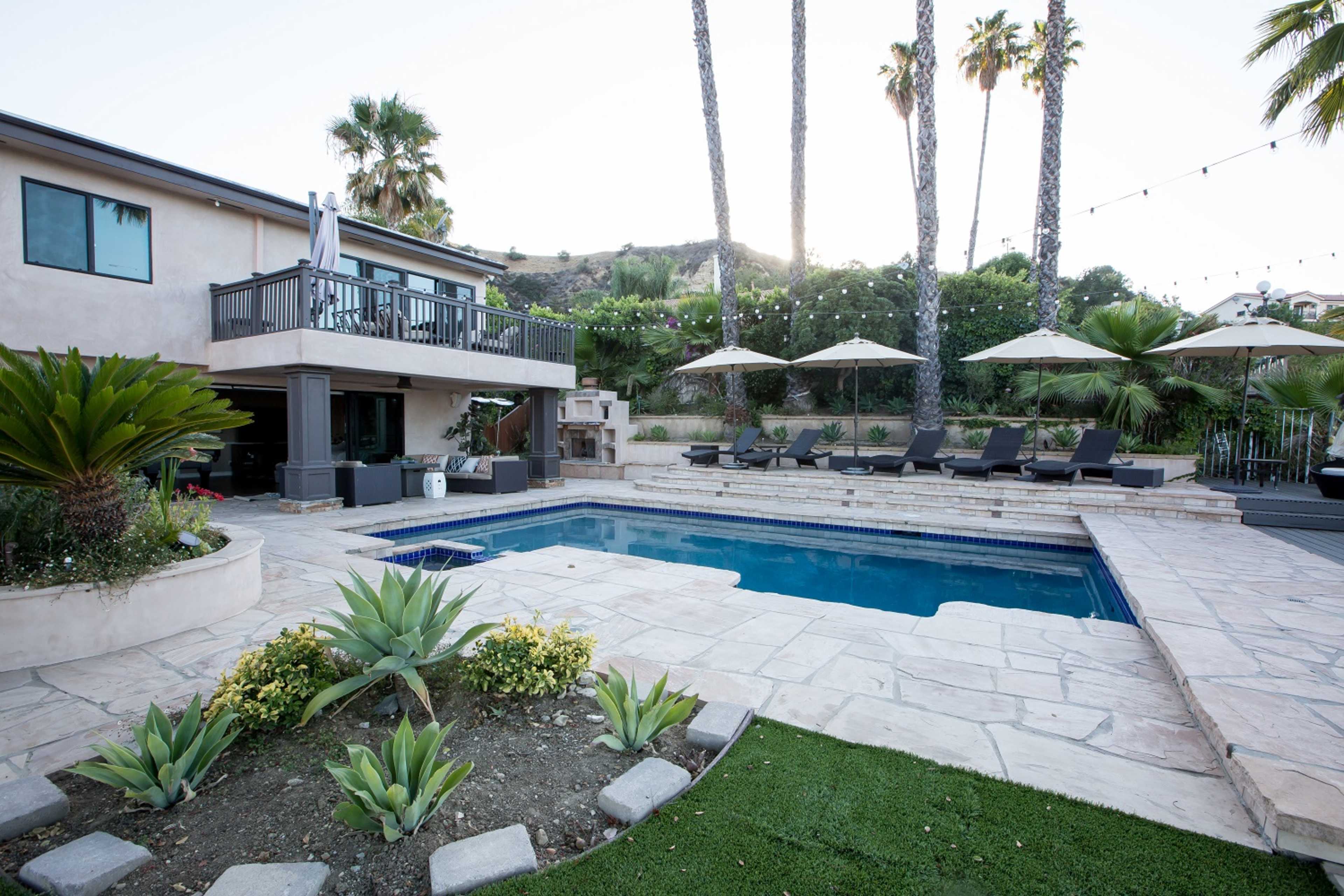 A backyard pool area featuring a landscaped design with lounge chairs, umbrellas, and palm trees surrounding a clear blue pool.