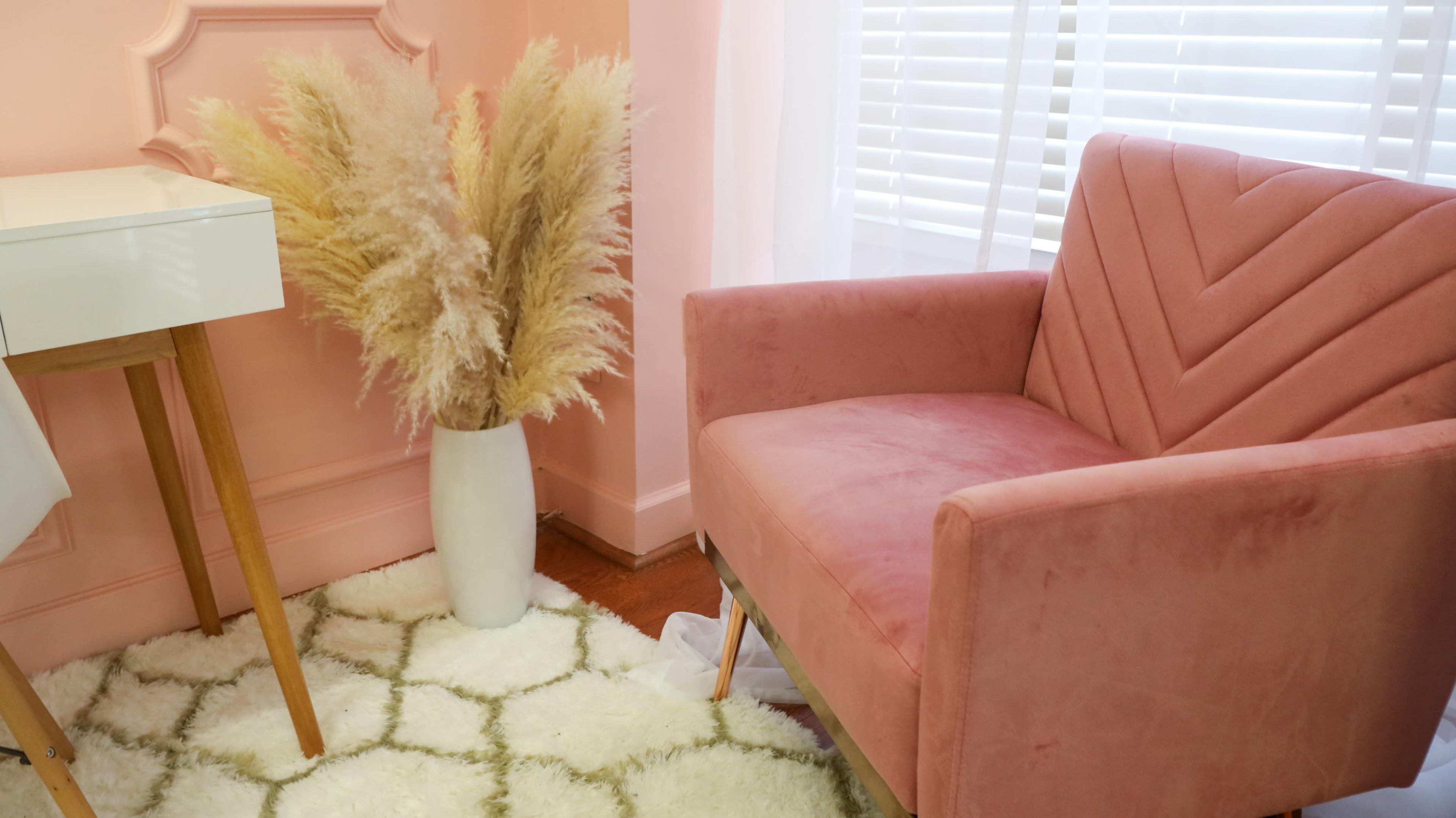 A pink upholstered armchair sits next to a white desk and a large vase of dried pampas grass on a textured rug in a light-colored room.