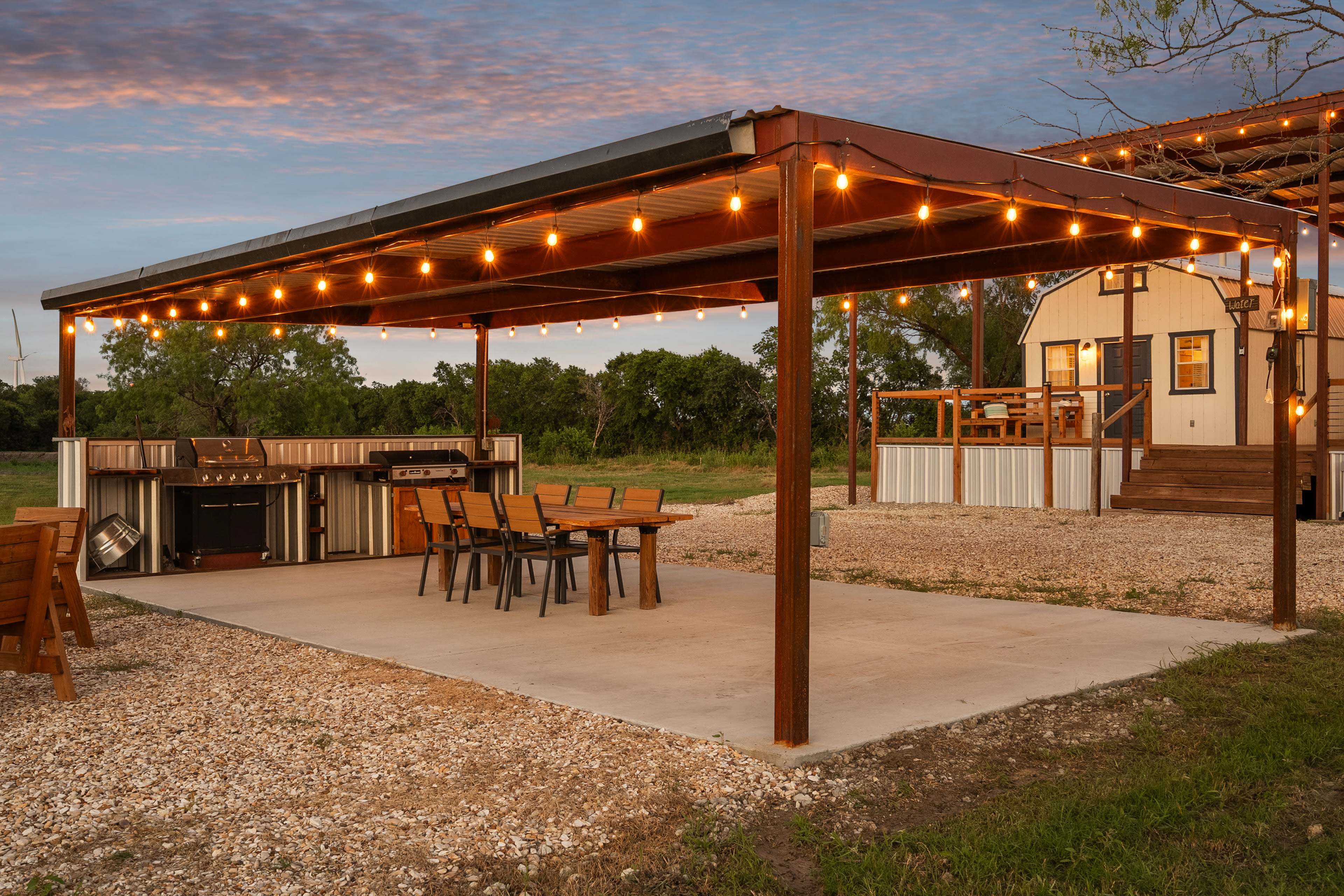 A covered outdoor area with a dining table, grill, and string lights is adjacent to a small building surrounded by greenery.