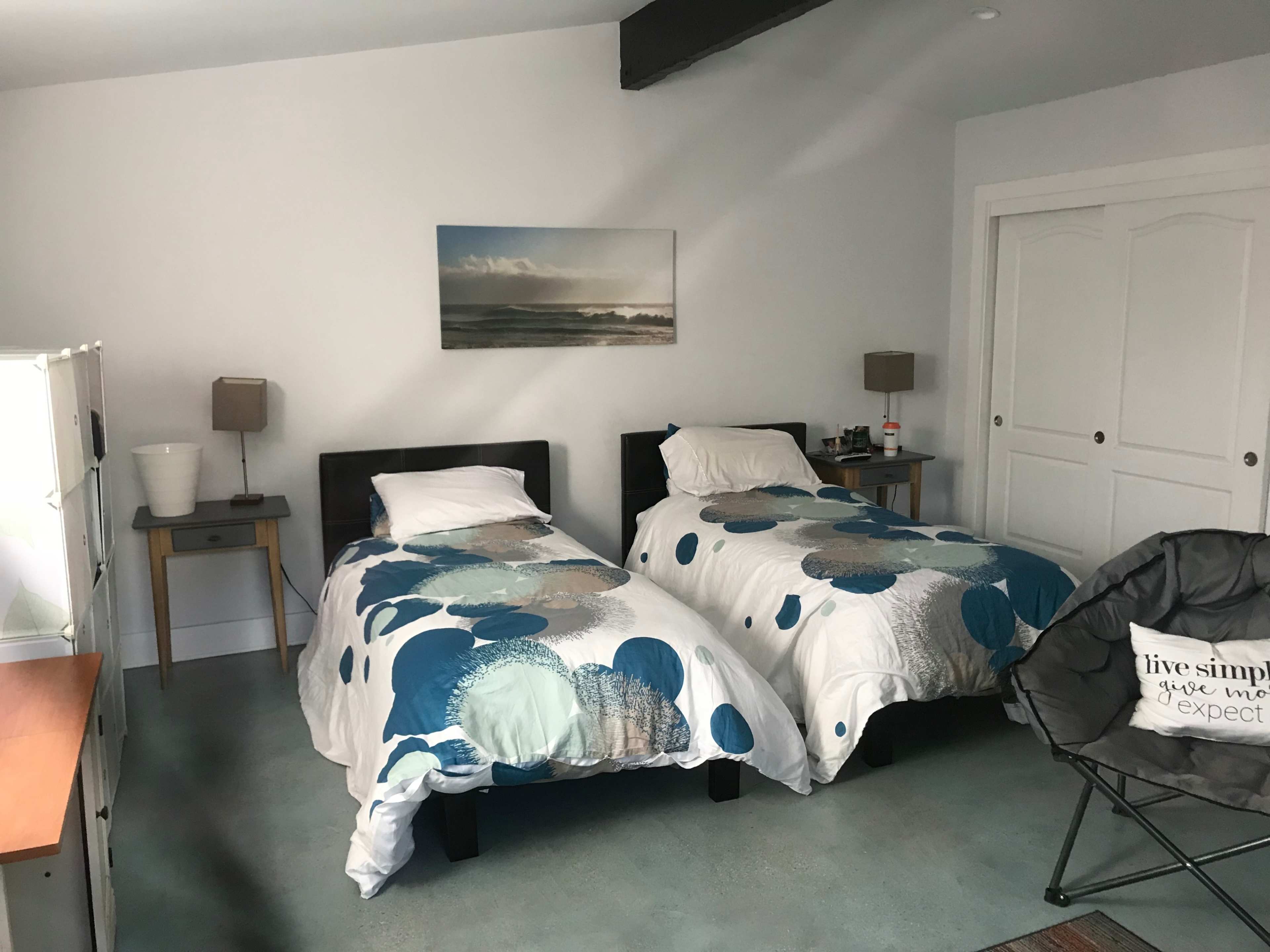 A bright bedroom featuring two twin beds with patterned bedding, a small dresser, and a comfortable chair in a minimalist style.