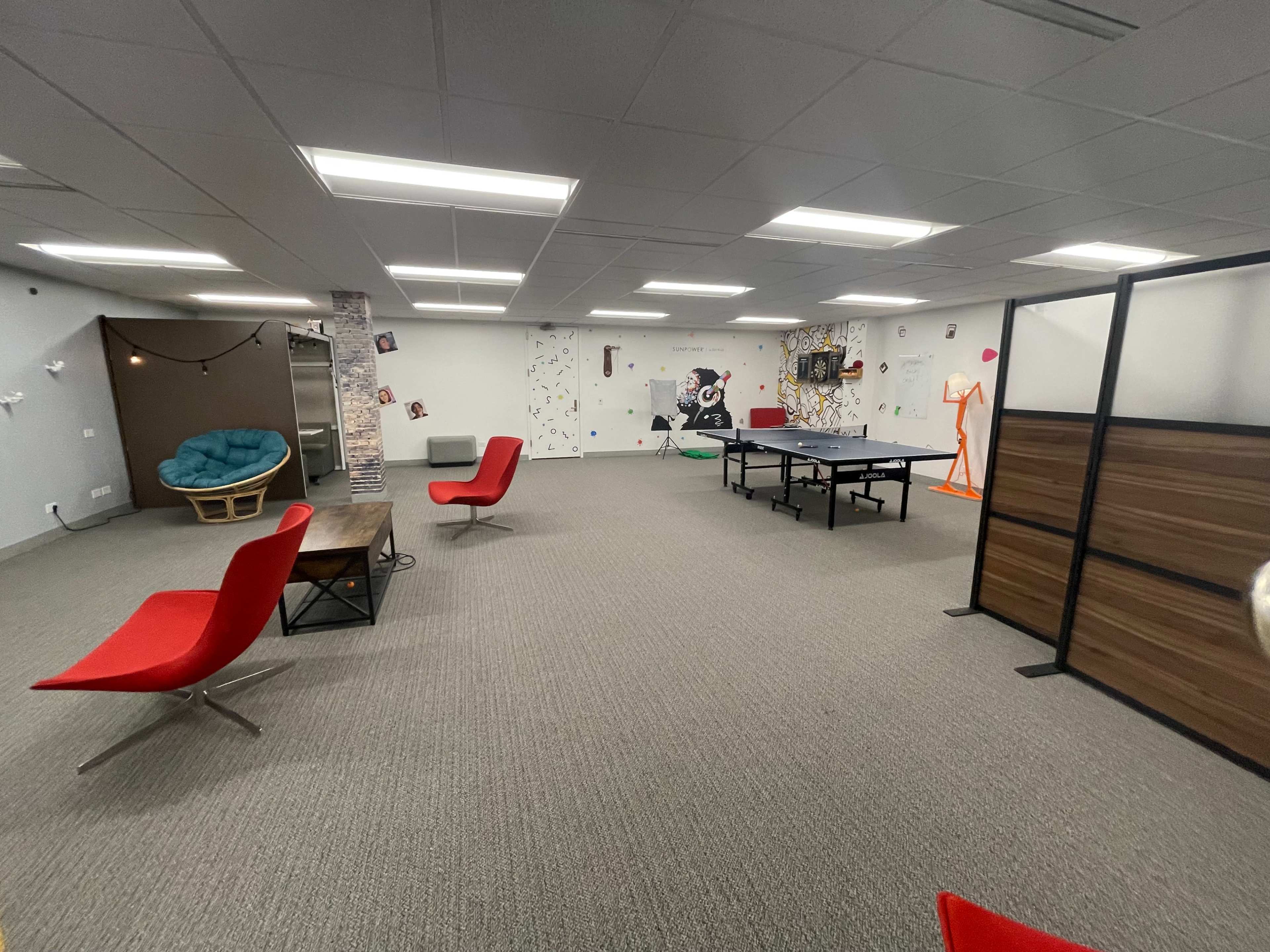 The image shows a spacious office with a ping pong table, colorful wall art, and modern furniture, including red chairs and a cozy seating area.