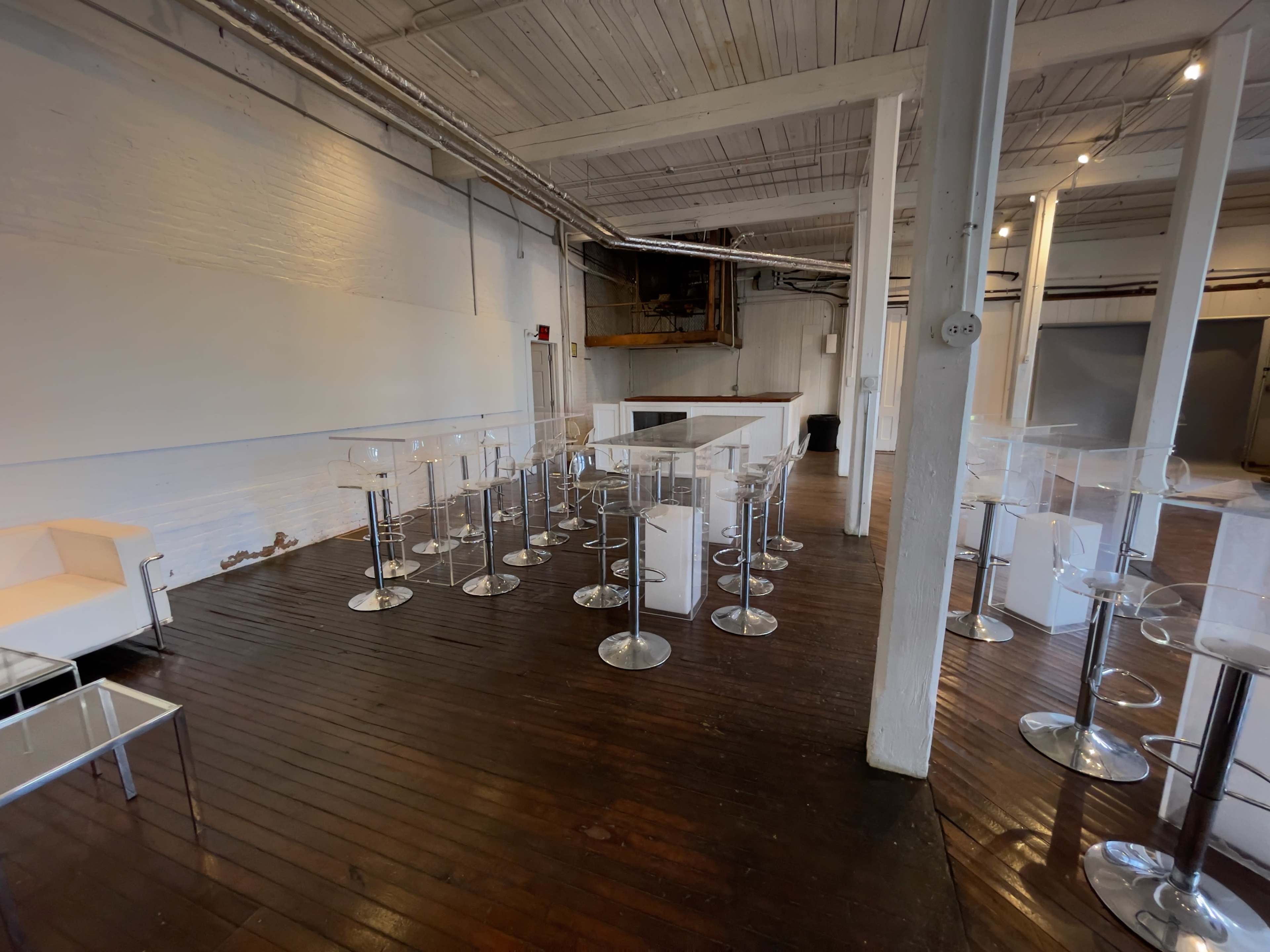 A spacious room with wooden floors, white walls, and clear high-top tables arranged throughout.
