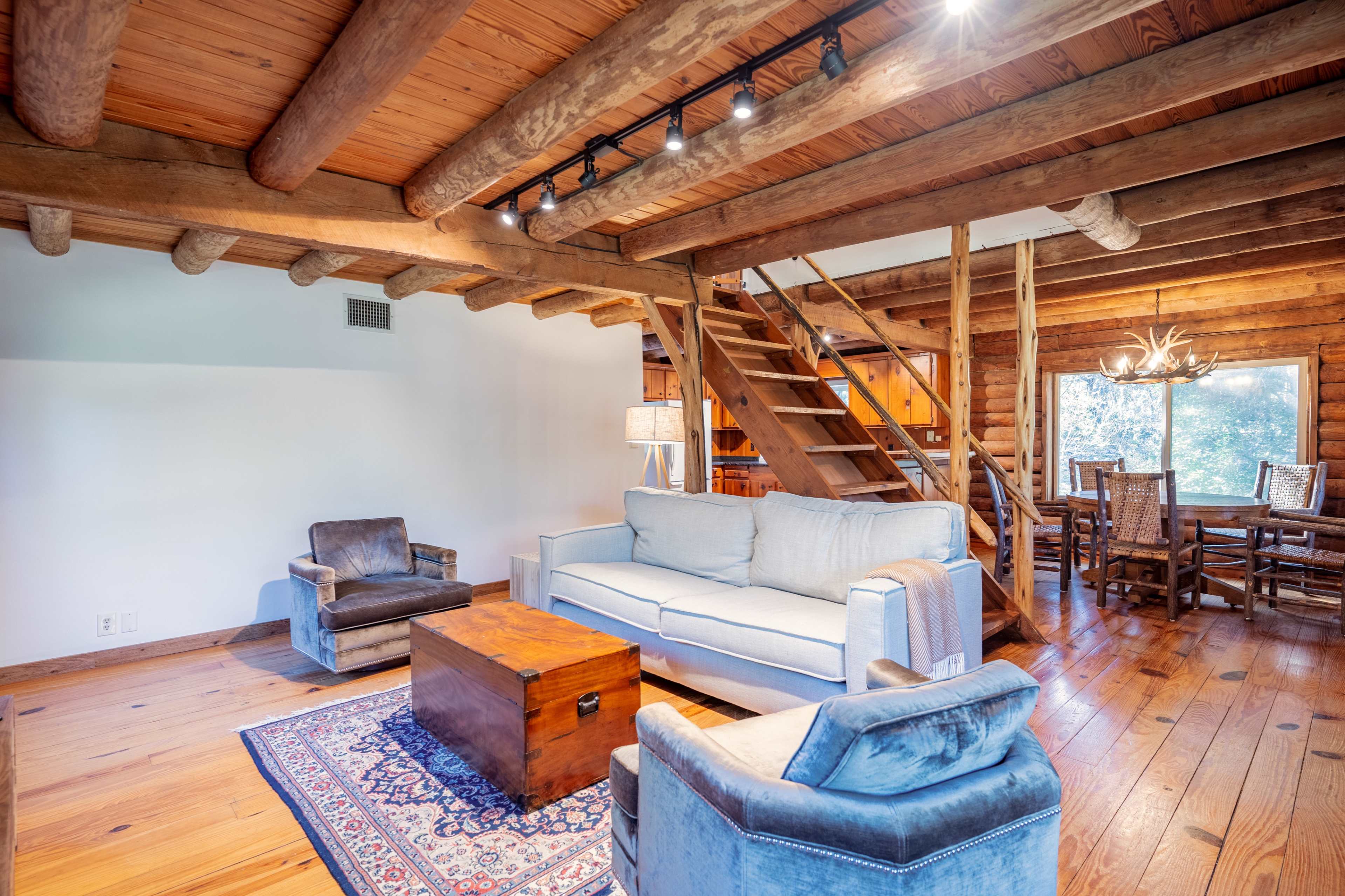 The image shows a rustic living room with wooden beams, a light-colored sofa, a wooden coffee table, and a staircase leading to an upper level.