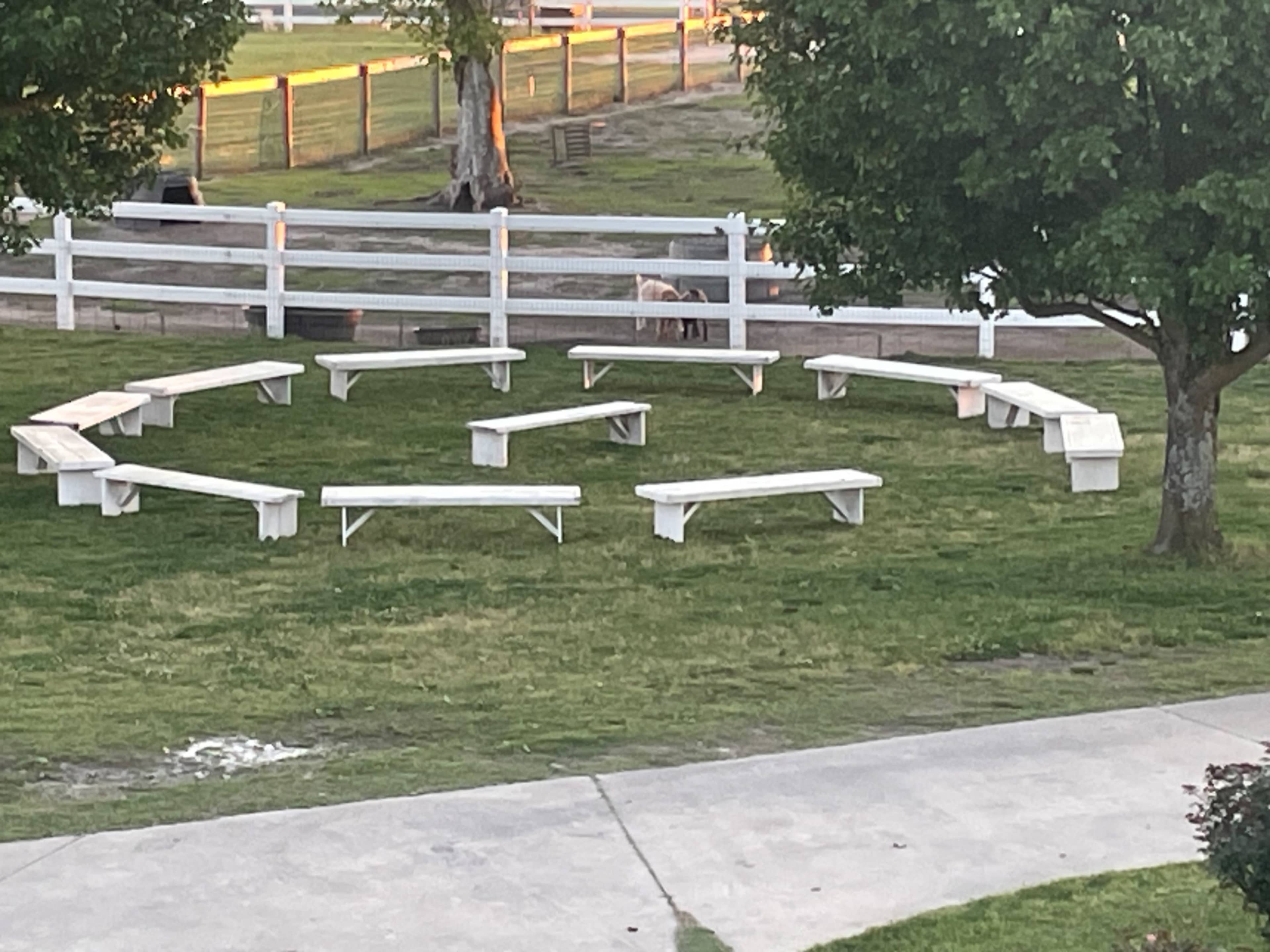 A circular arrangement of white benches on a grassy area near a wooden fence.