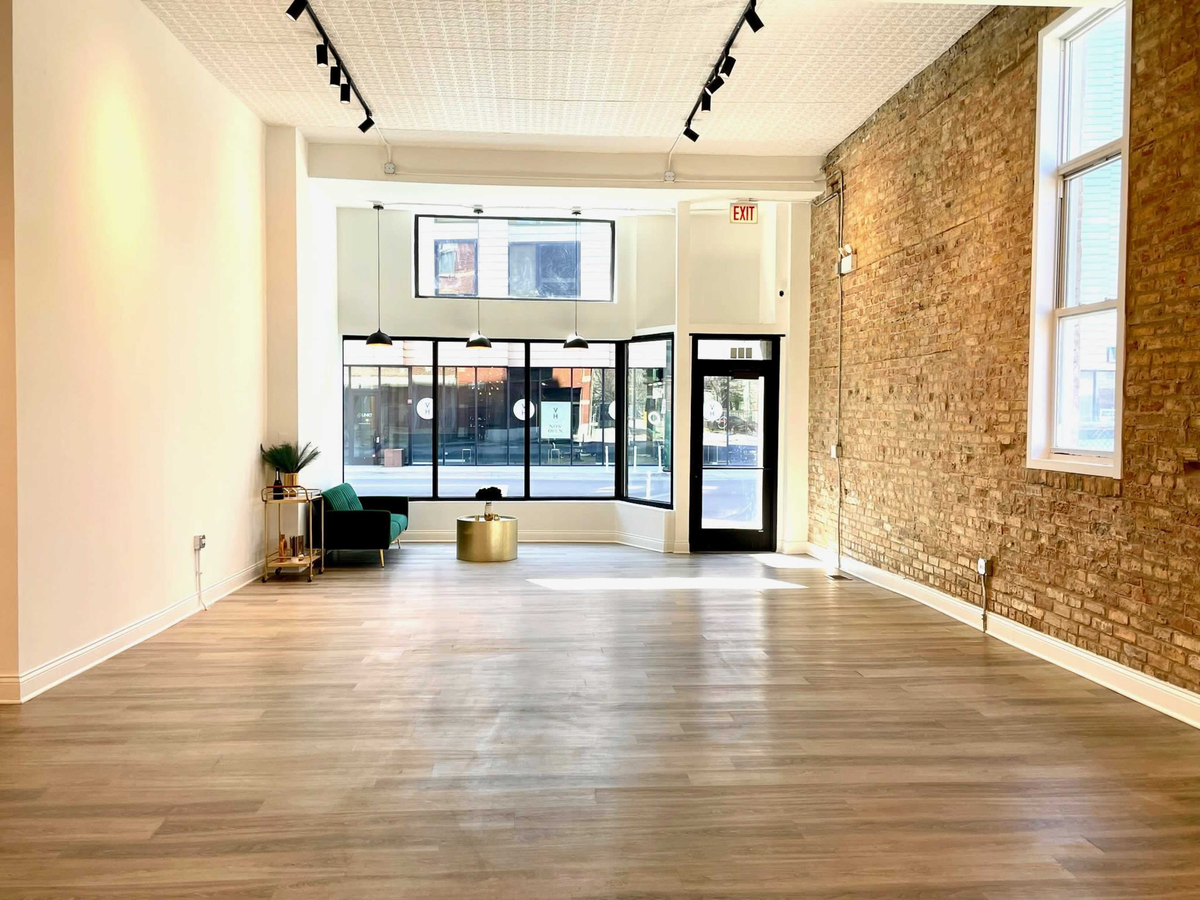 An empty, well-lit interior space with wooden flooring, large front windows, and exposed brick walls.