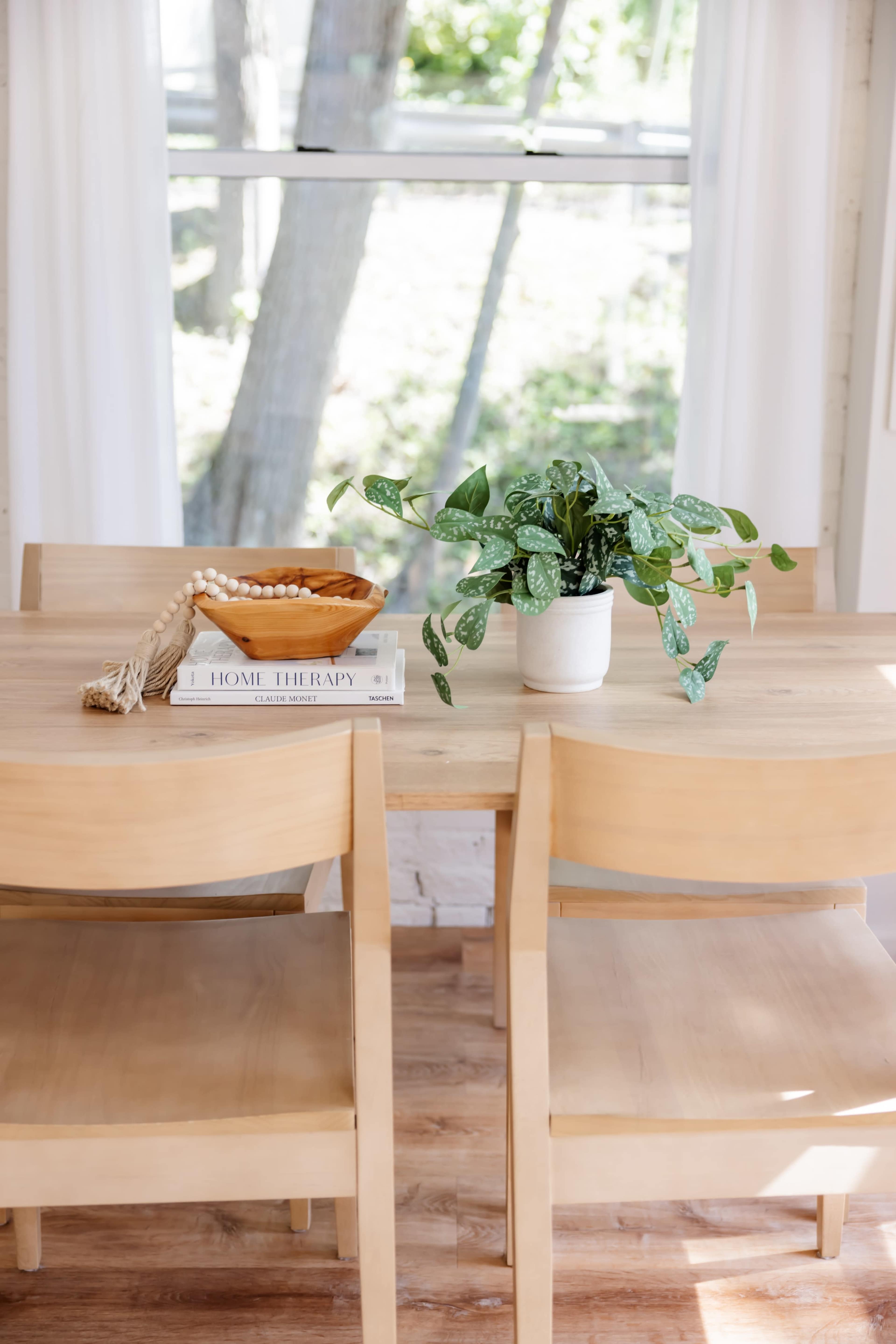 A wooden dining table is set with a potted plant, a decorative bowl, and a stack of books, with sunlight filtering through the windows behind it.
