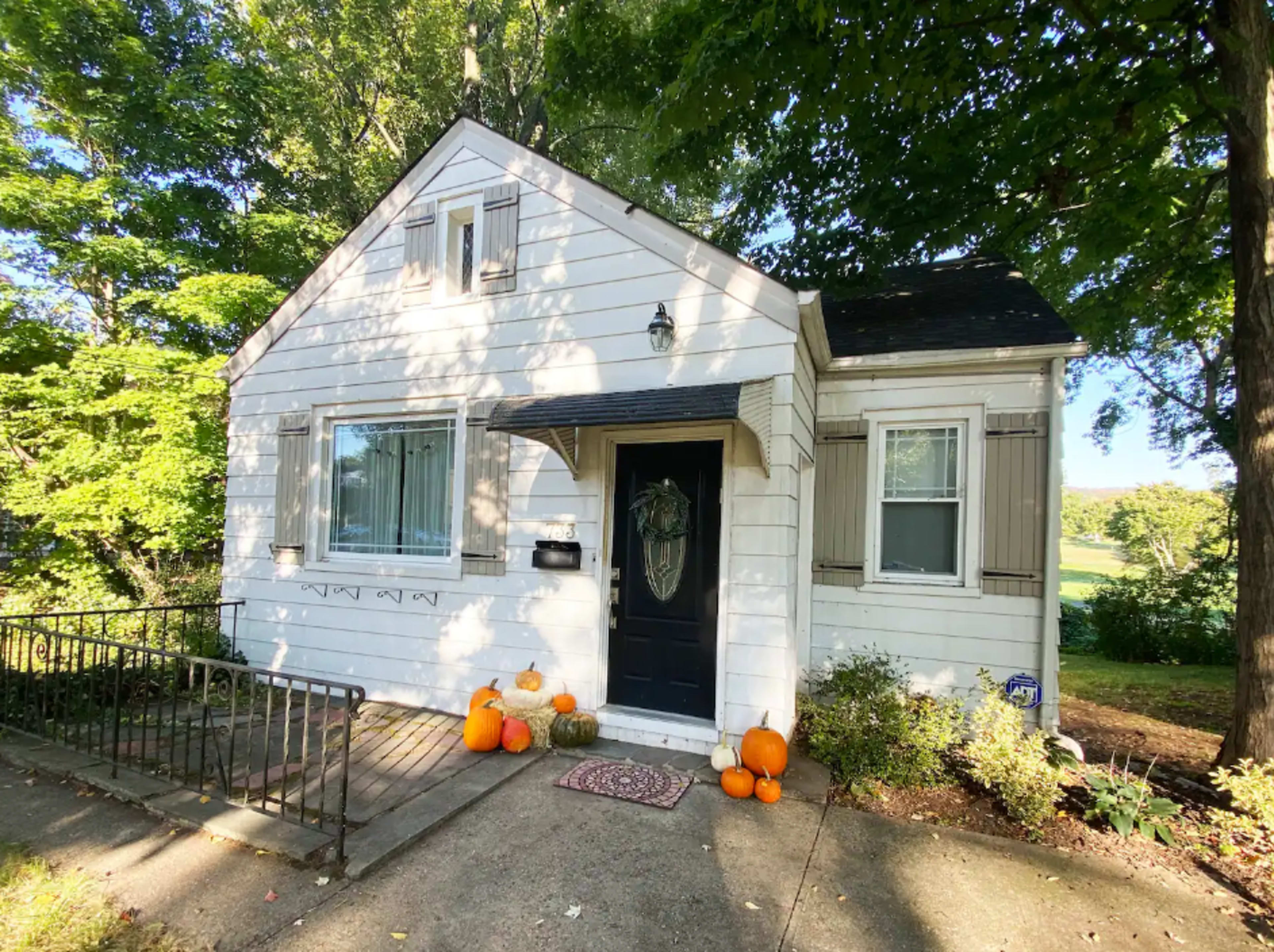 A small white house with a black door has pumpkins arranged on the porch and is surrounded by trees.