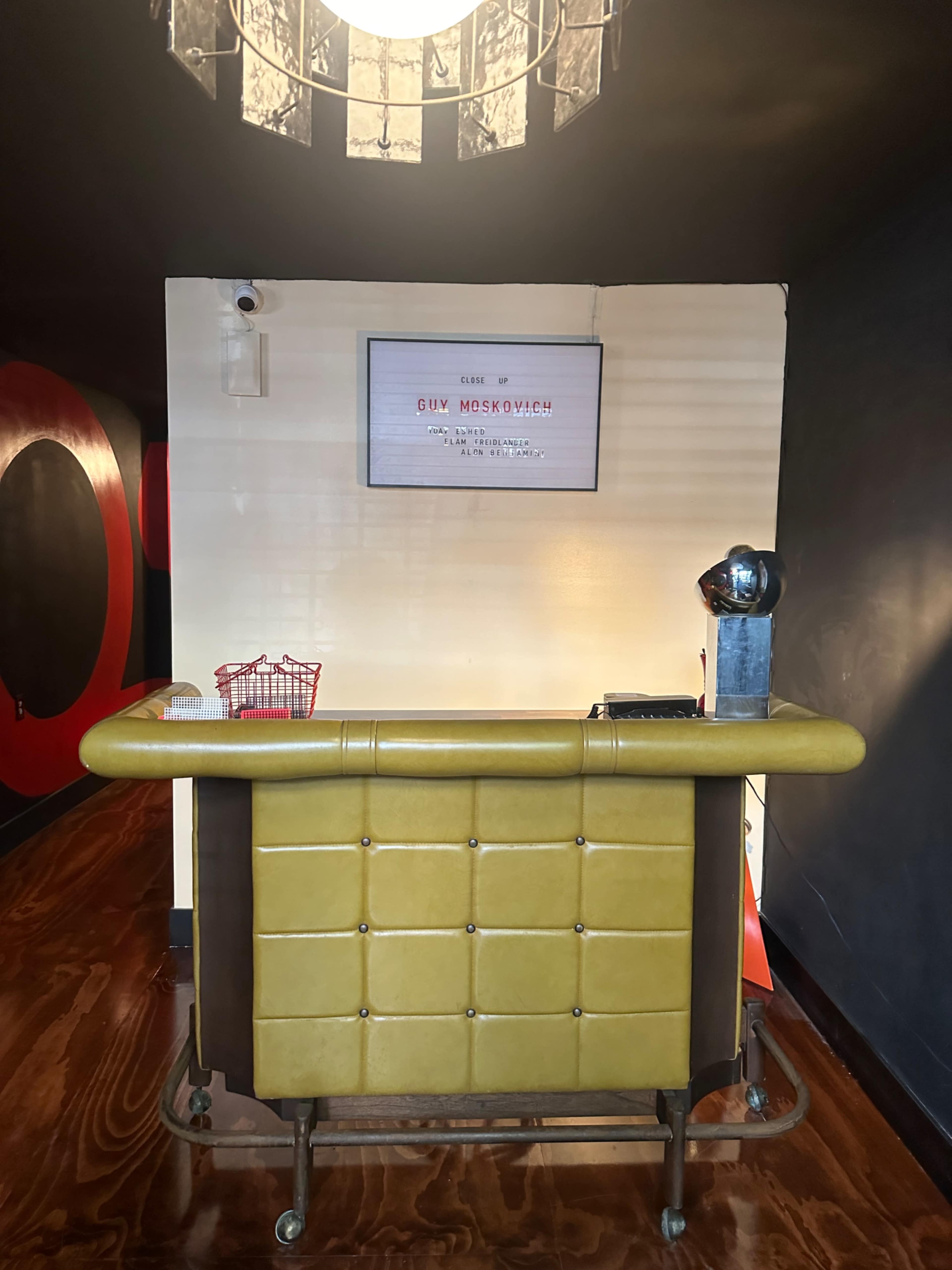 The image shows a reception desk with a yellow front, a decorative wall-mounted sign, and a vintage-style lamp above.