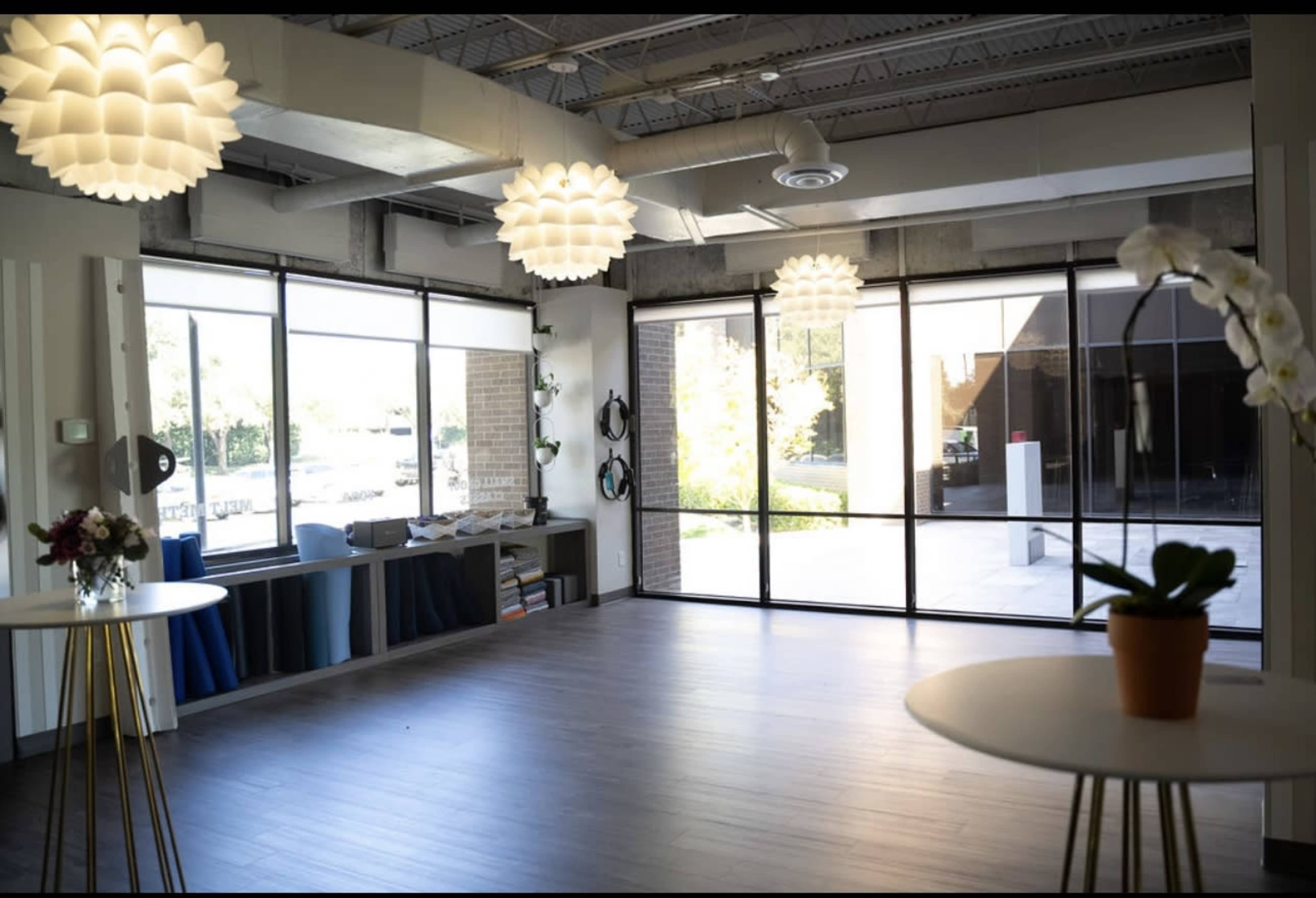 A well-lit interior space features large windows, modern light fixtures, and small round tables with potted plants.