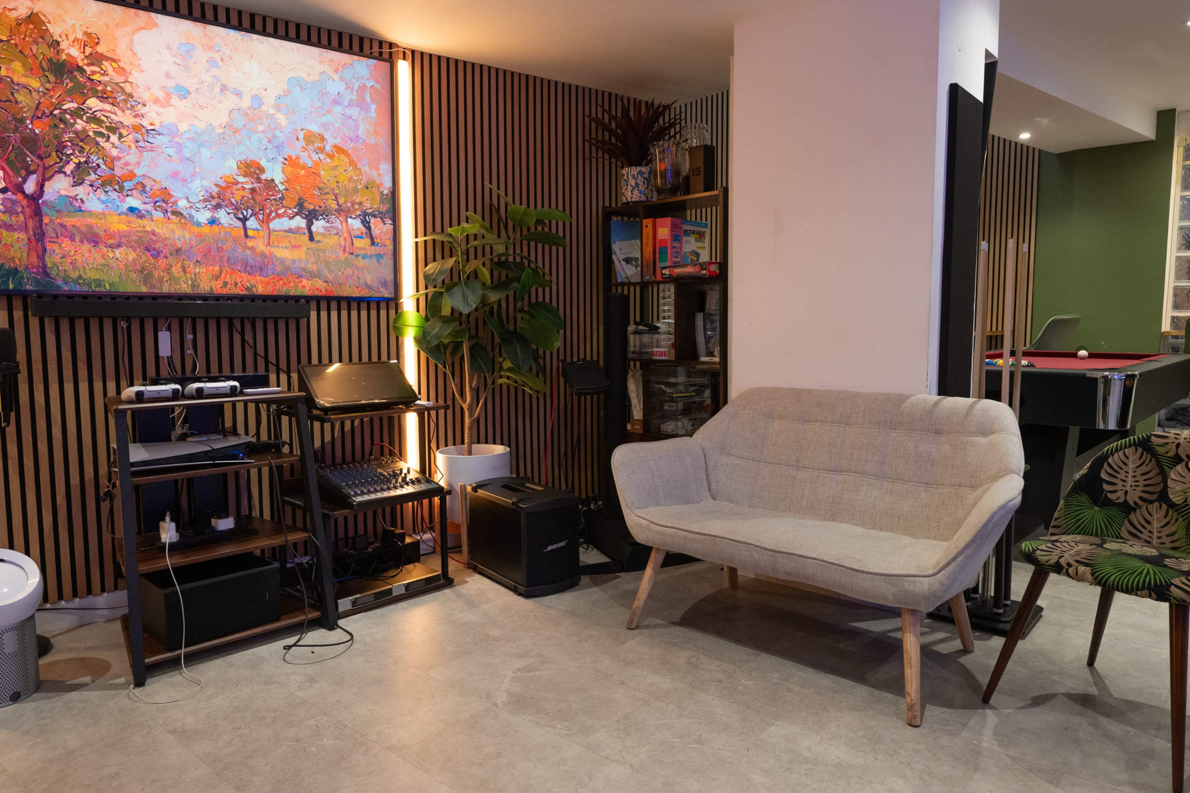 The image shows a cozy interior space with a light-colored couch, a wooden shelving unit filled with electronics and greenery, and a colorful painting on the wall.