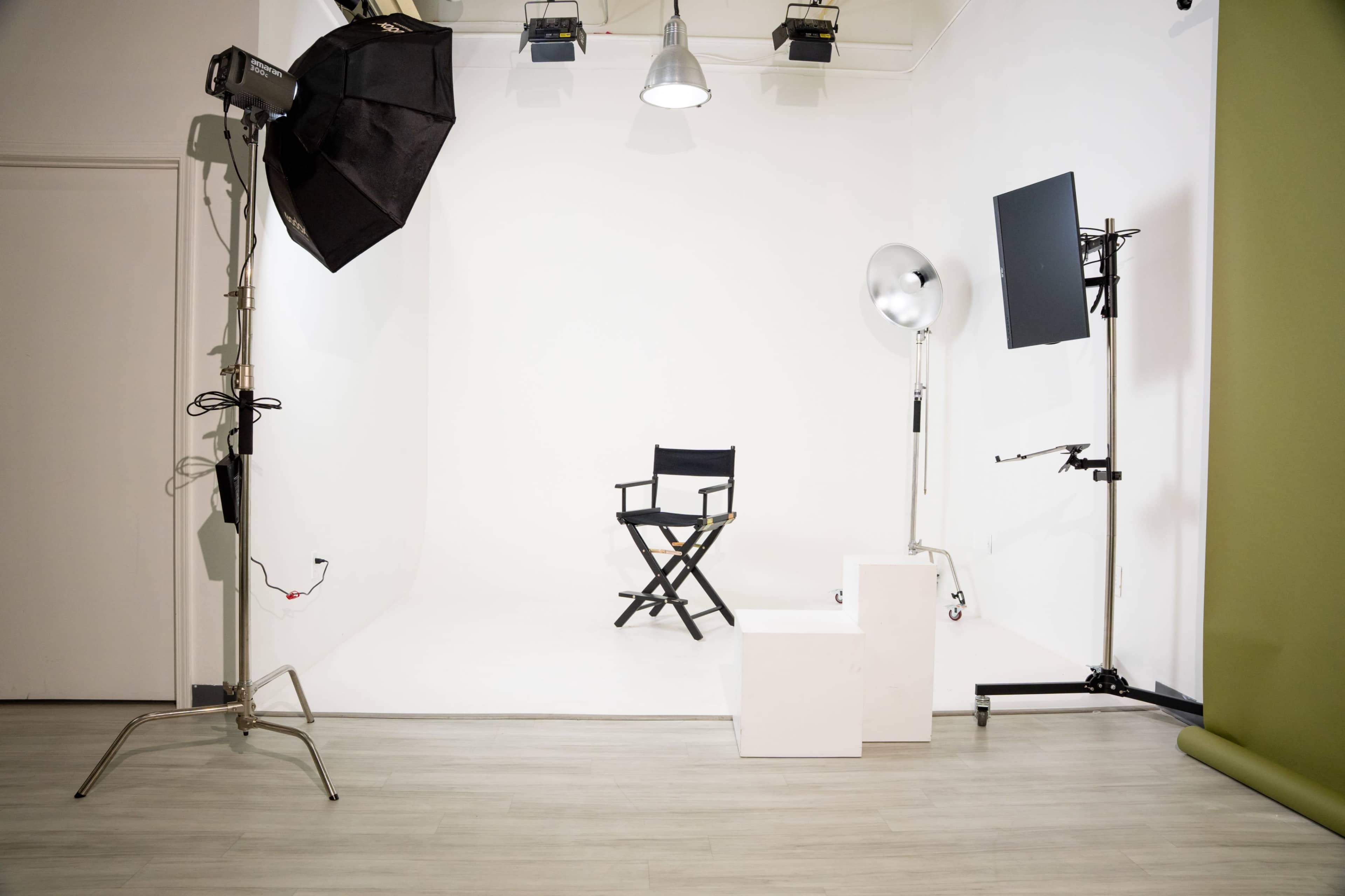 The image shows a minimalist photography studio setup featuring a director's chair, lighting equipment, and a blank backdrop.
