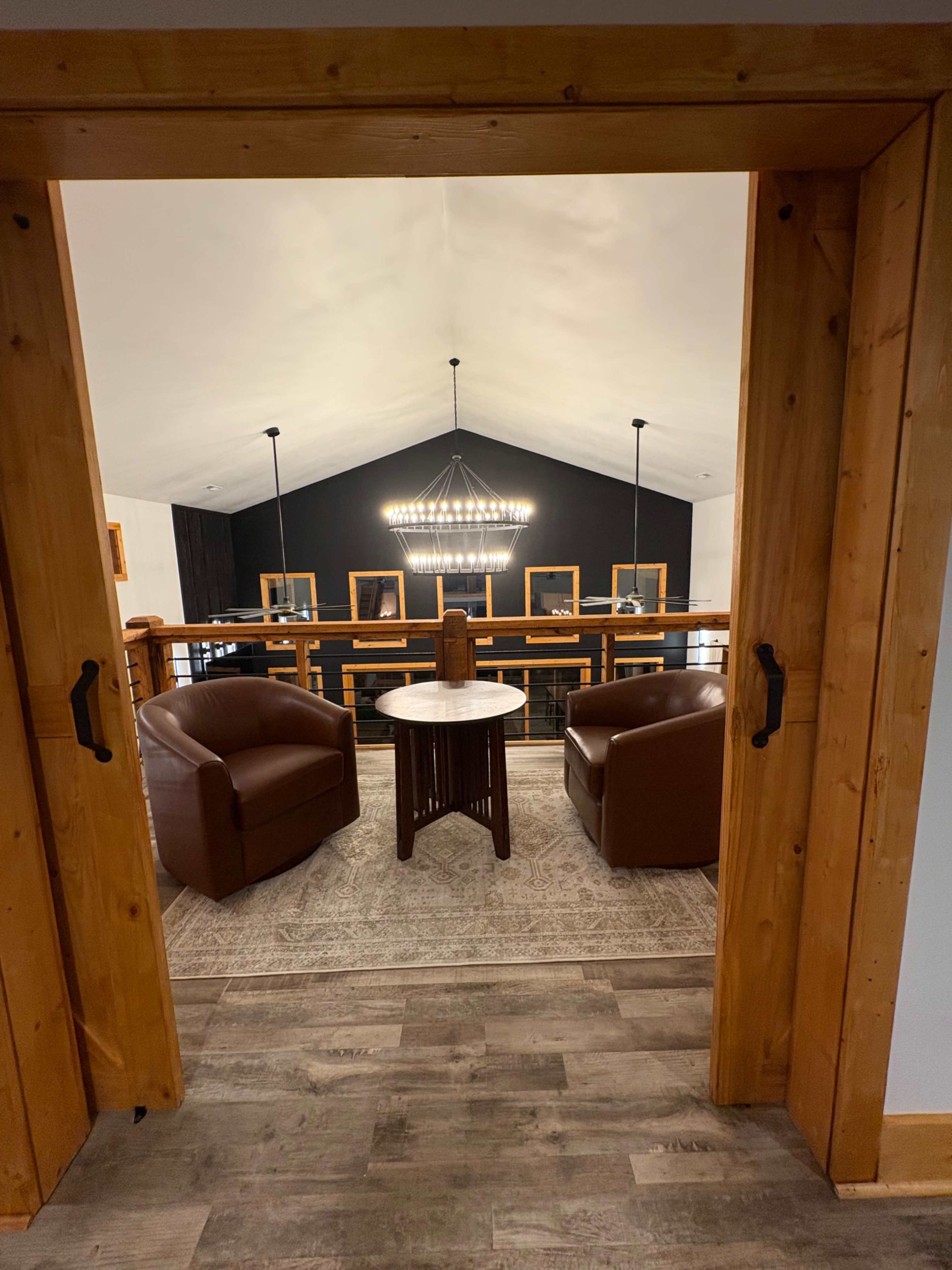 The image shows a view from a doorway into a loft area featuring two brown leather chairs, a small round table, and a chandelier above, with framed pictures on the walls.
