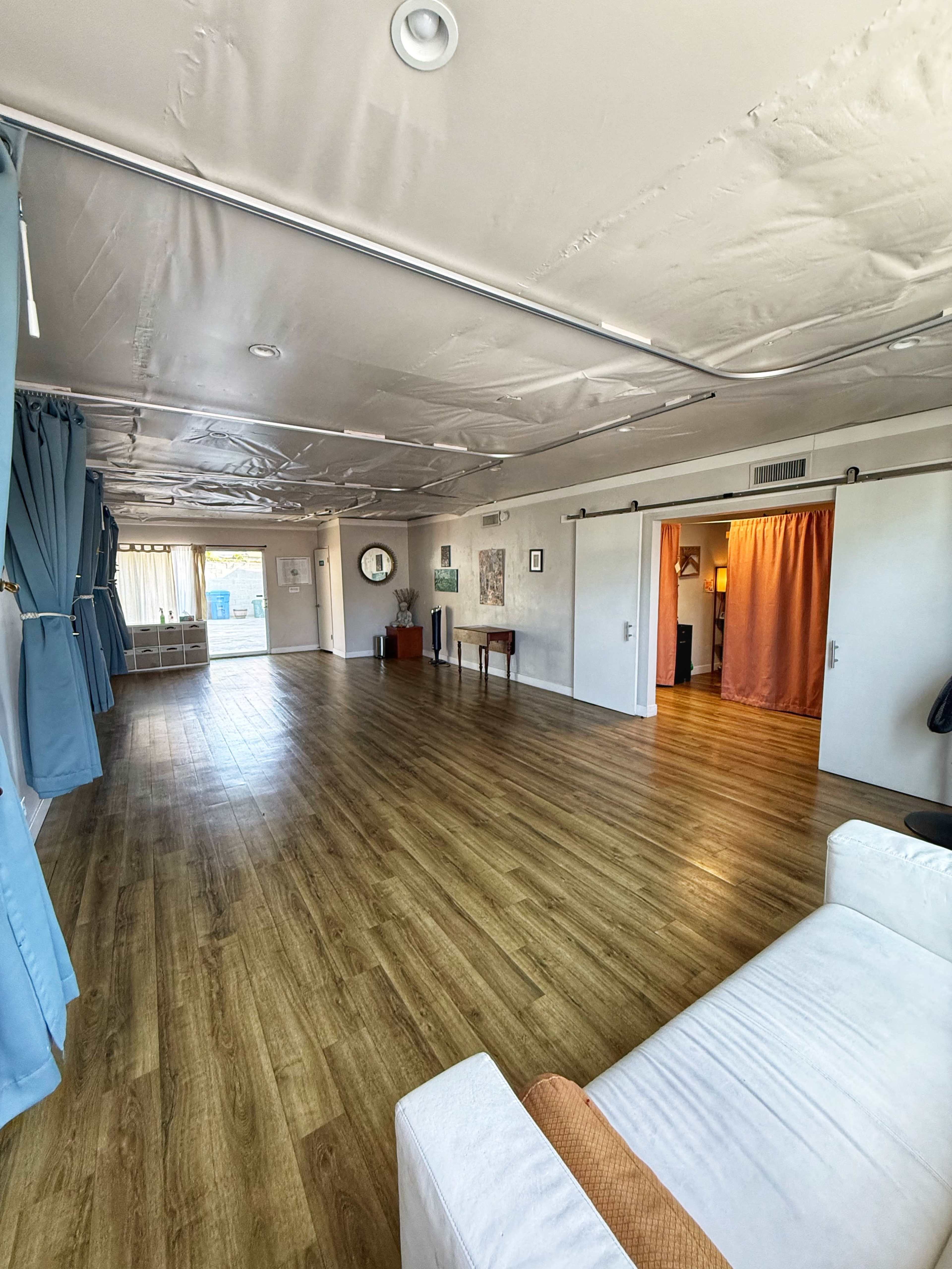 The image shows a spacious, illuminated room with wooden flooring, featuring a cozy seating area and two curtains leading to separate spaces.