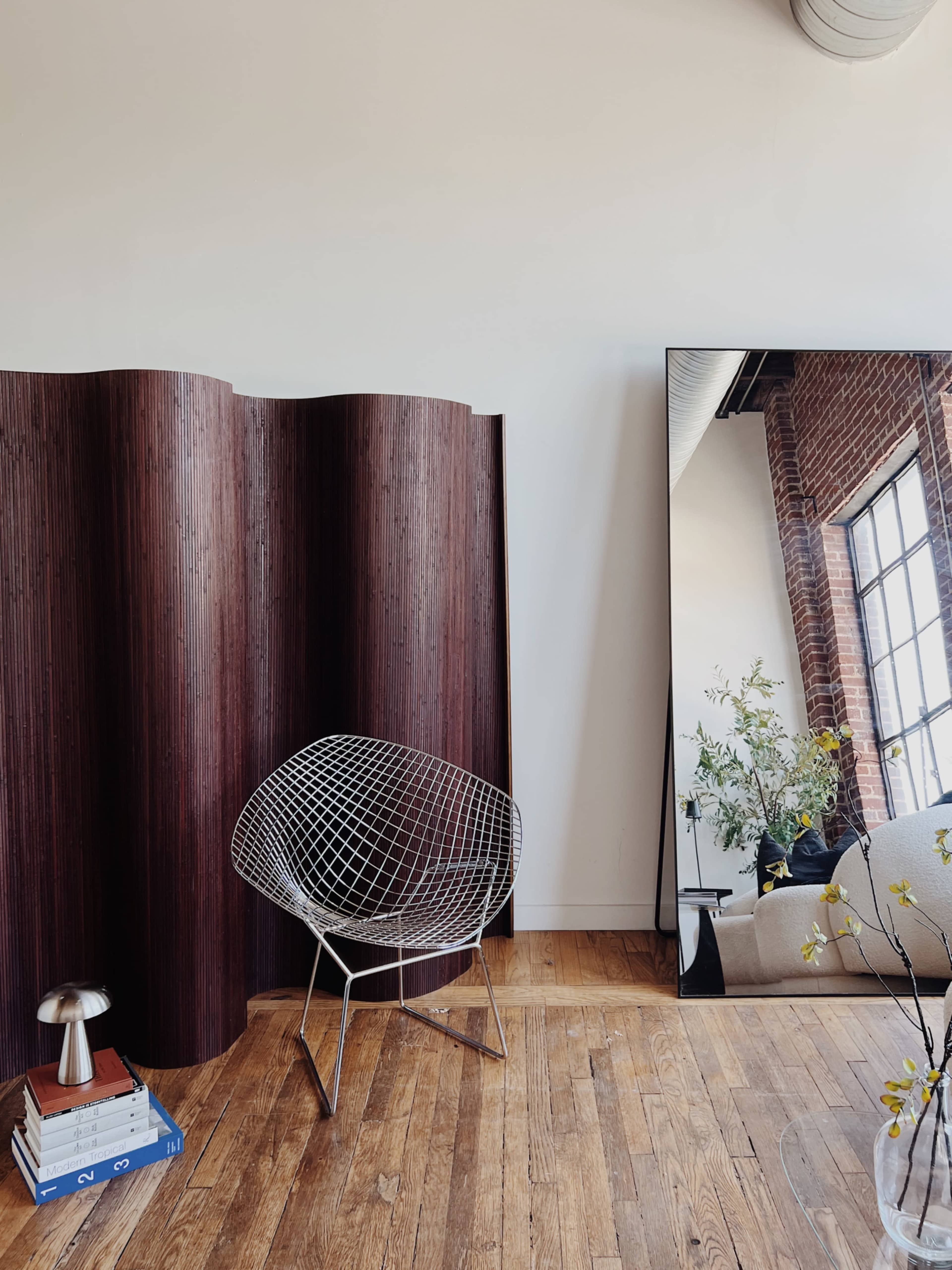 The image features a modern chair made of wire beside a curved wooden partition and a large mirror reflecting a bright, open space with a couch and a window.