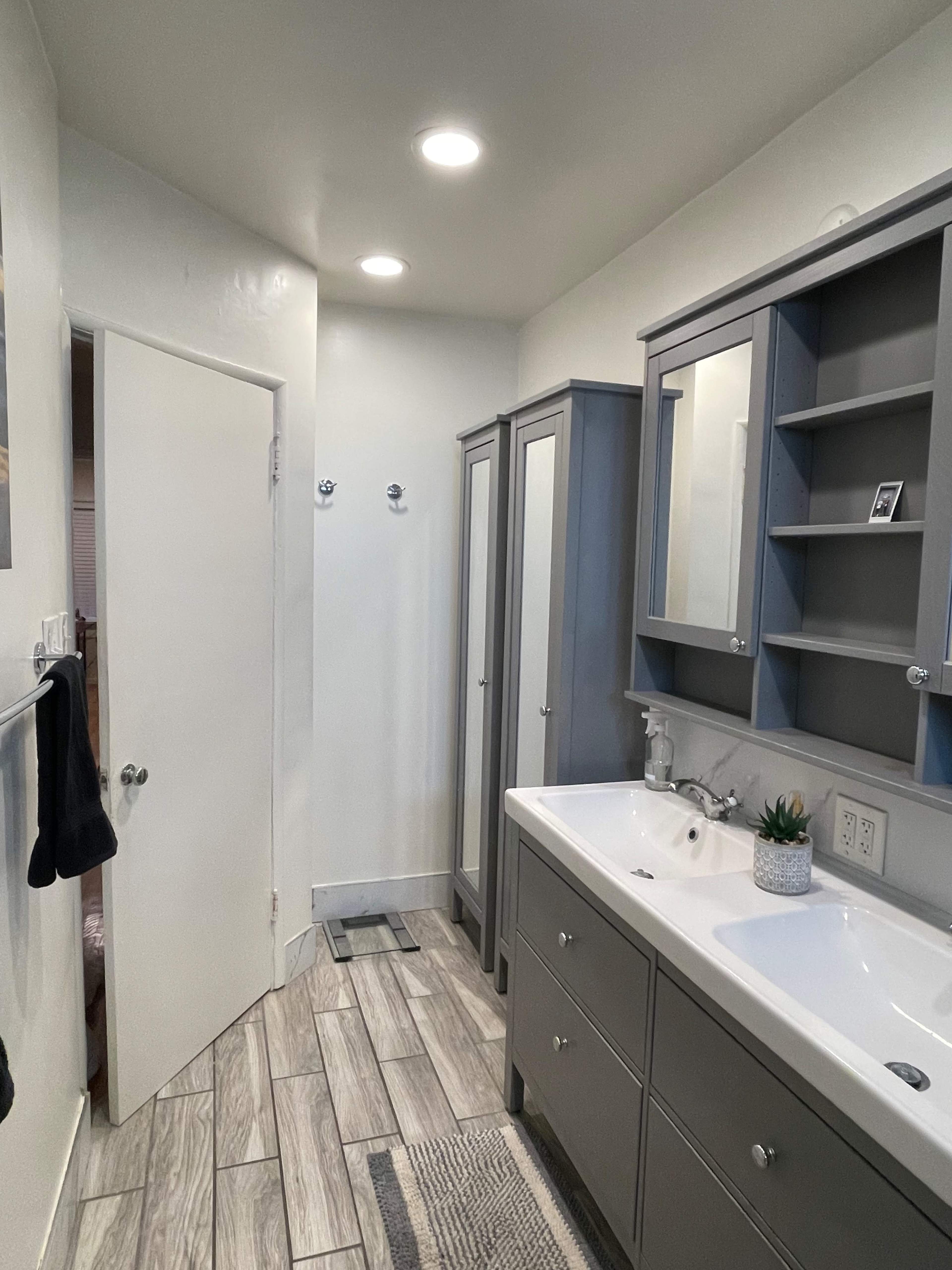 The image shows a modern bathroom with gray cabinets, a double sink vanity, and a door leading to another area.