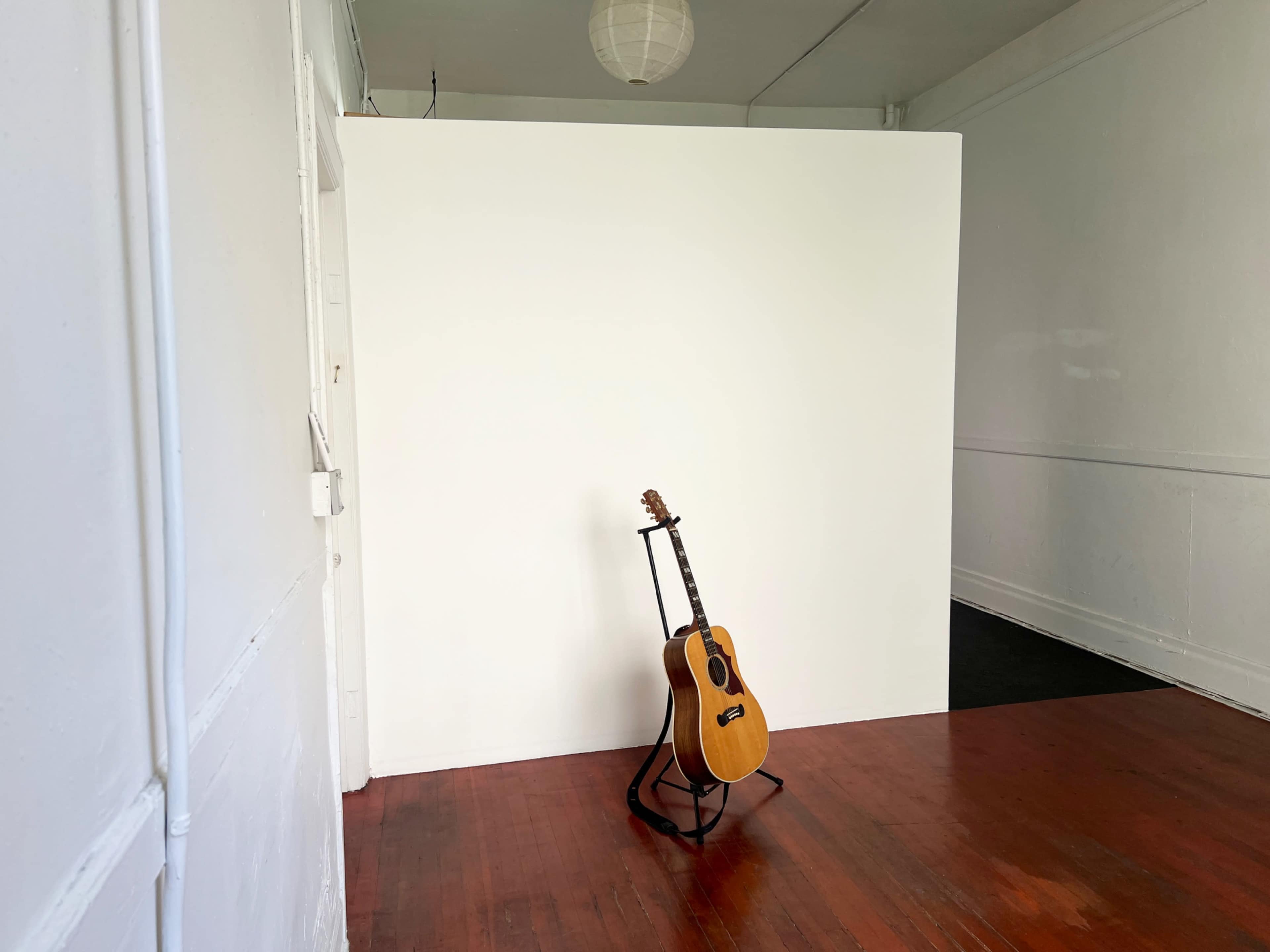 A wooden guitar stands on a stand in front of a plain white wall in a room with hardwood flooring.