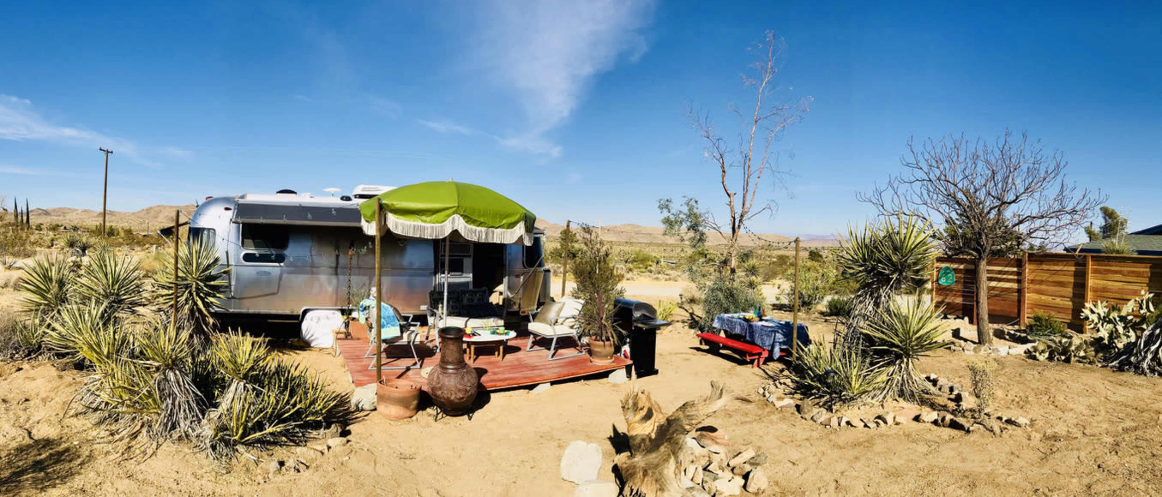 A silver Airstream trailer is parked on a wooden deck surrounded by desert vegetation and a few outdoor furnishings under a green awning.
